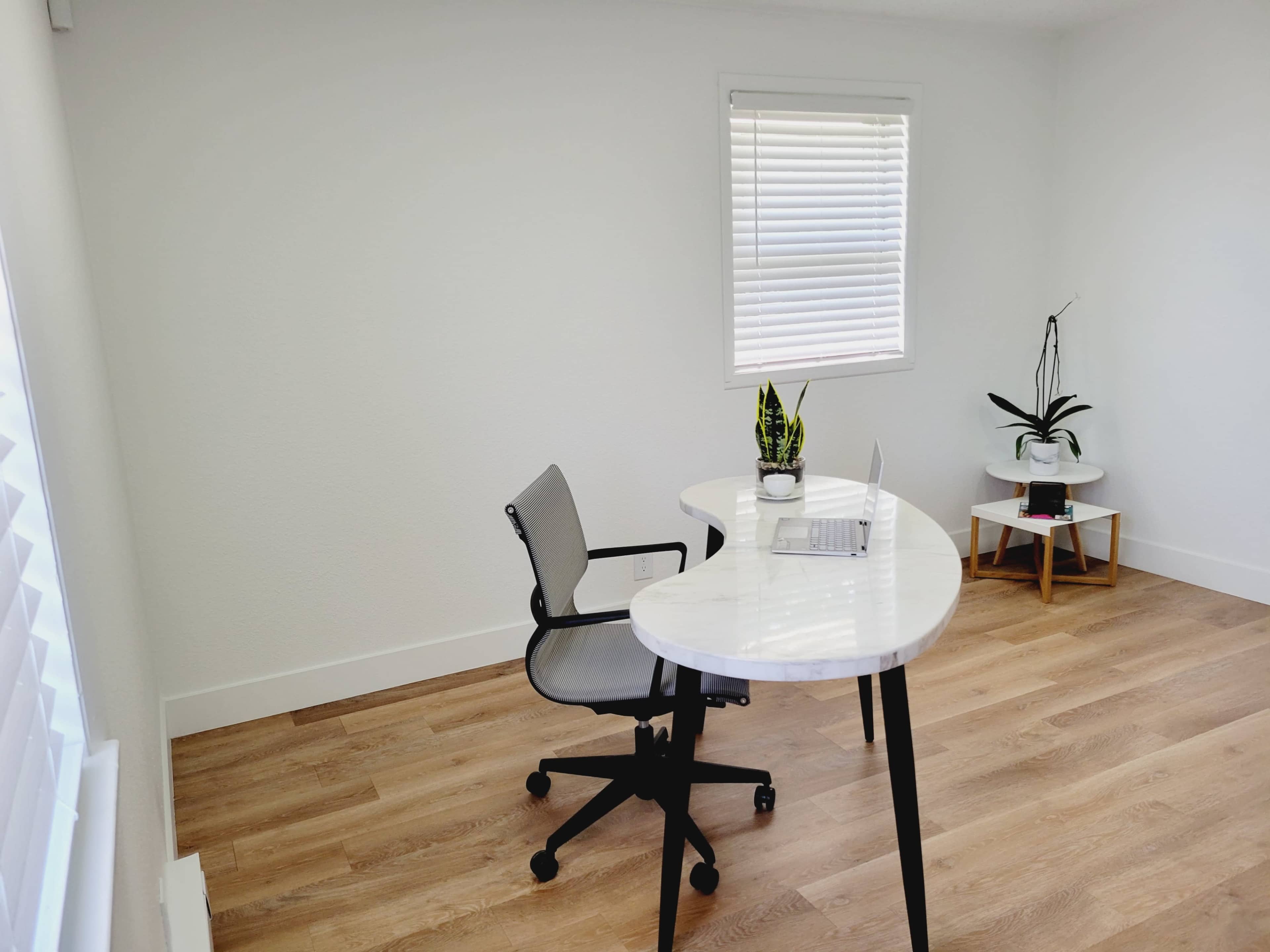 A modern workspace features a white desk with a laptop, a grey office chair, and potted plants, set against a light-colored wall and hardwood floor.