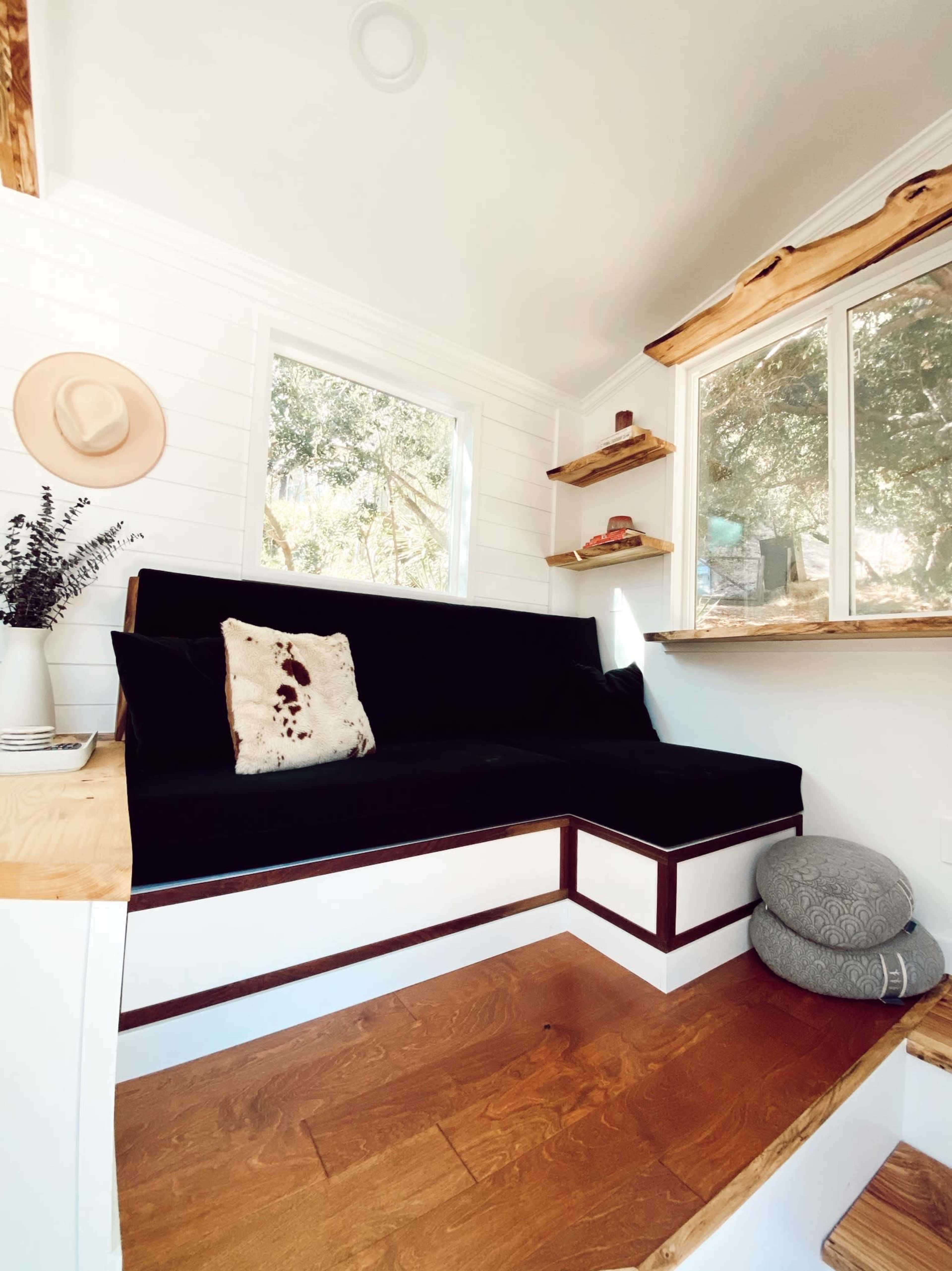 The image shows a cozy interior space with a black couch, wooden steps, and large windows, illuminated by natural light.