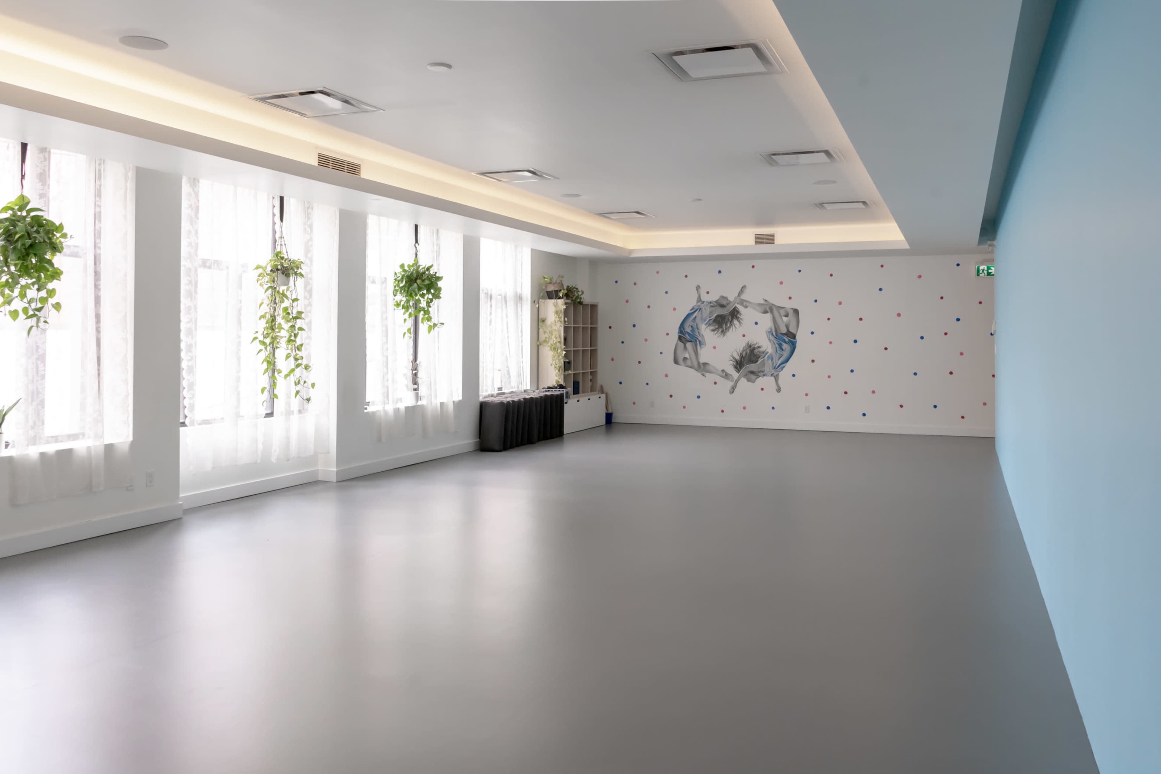 A spacious, well-lit room with large windows featuring indoor plants, a simple blue wall, and a mural on one side.
