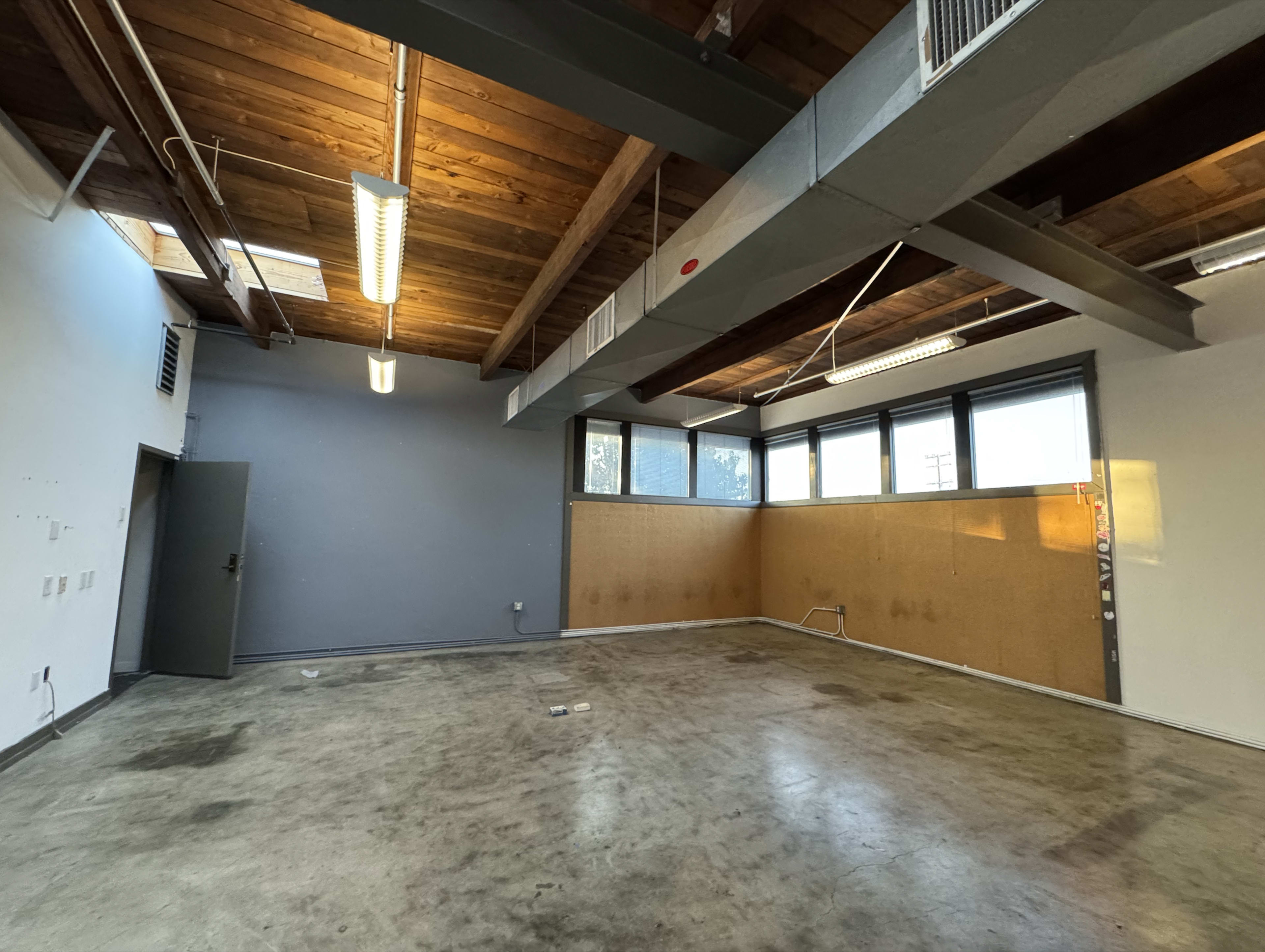 The image shows a spacious, unfinished room with exposed wooden beams and a concrete floor, featuring large windows along one wall.