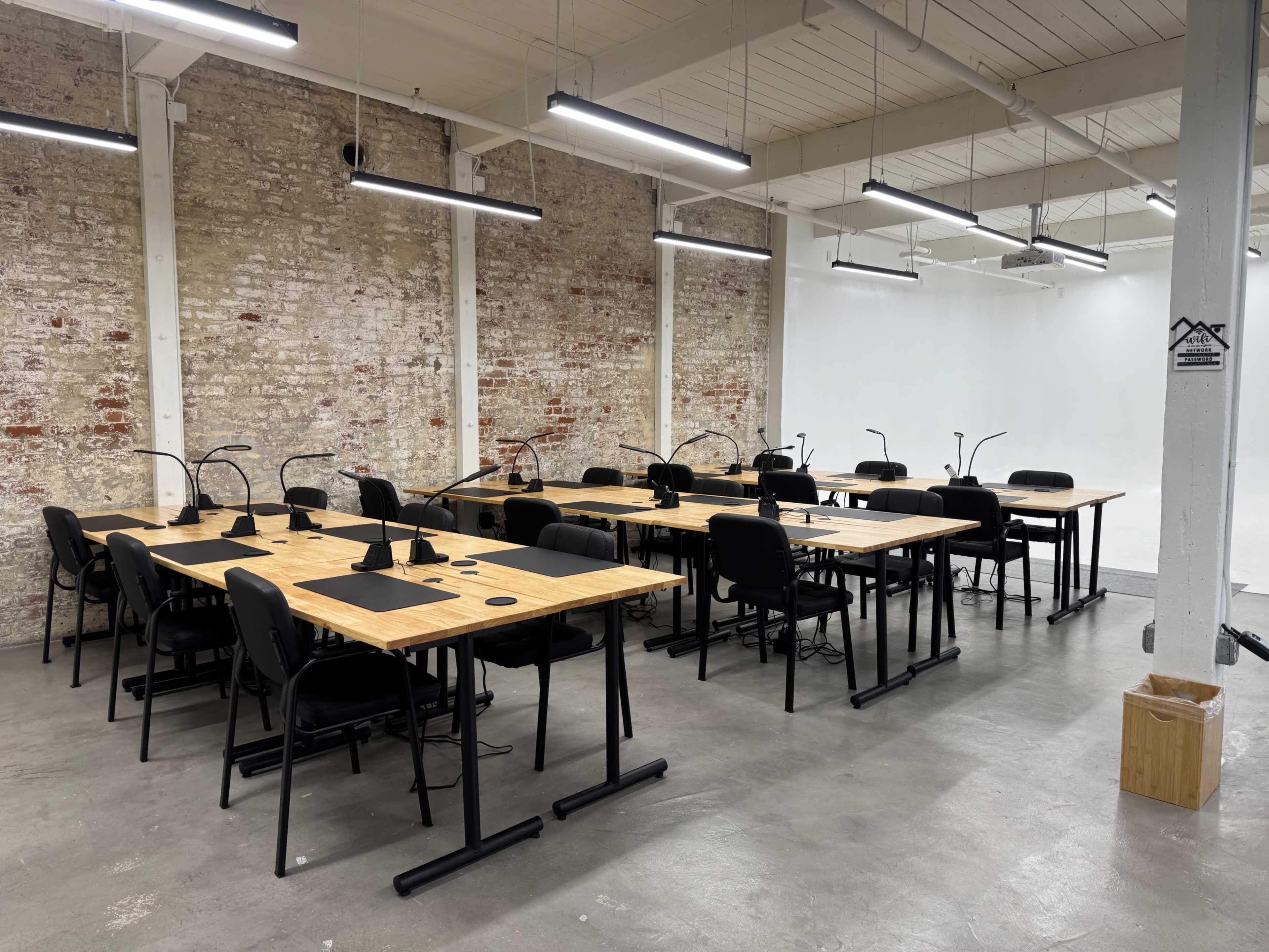 The image shows a spacious room with several wooden desks and black chairs arranged in rows against a brick wall.