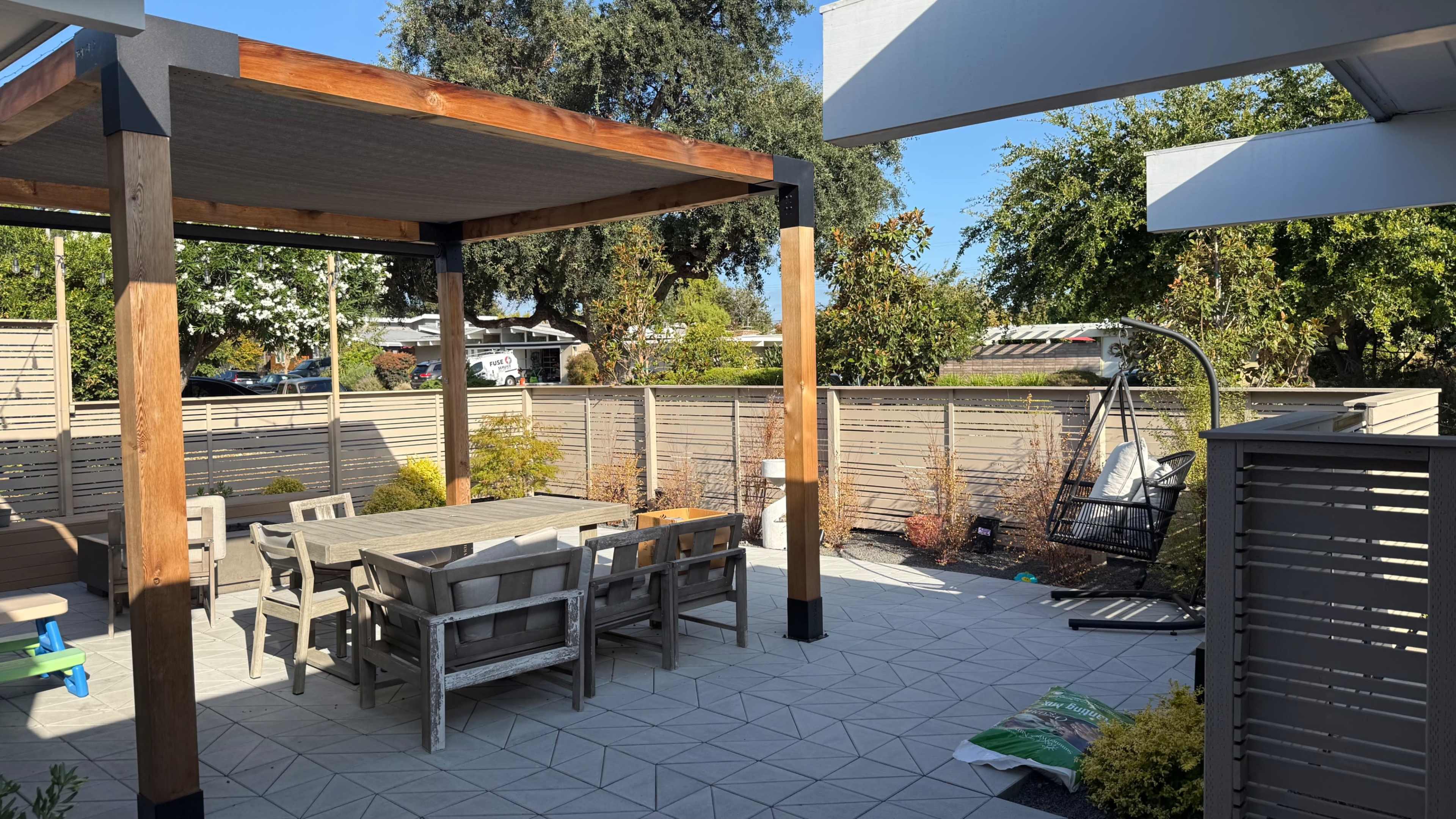 The image shows a backyard patio with a wooden gazebo, a rectangular dining table, and a swing chair surrounded by low fences and landscaping.
