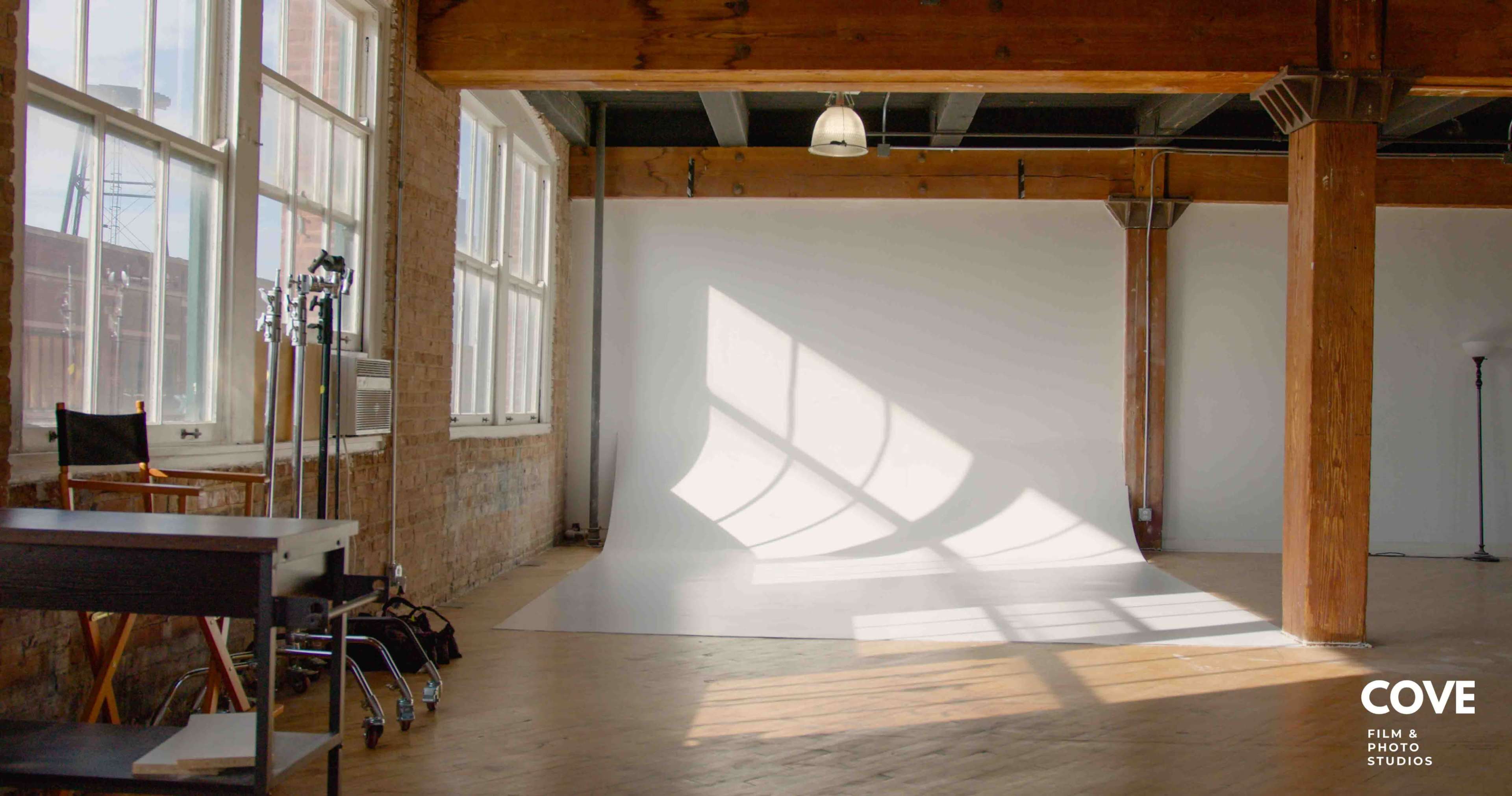 A well-lit photo studio features a white backdrop, wooden floors, and large windows casting shadows on the floor.