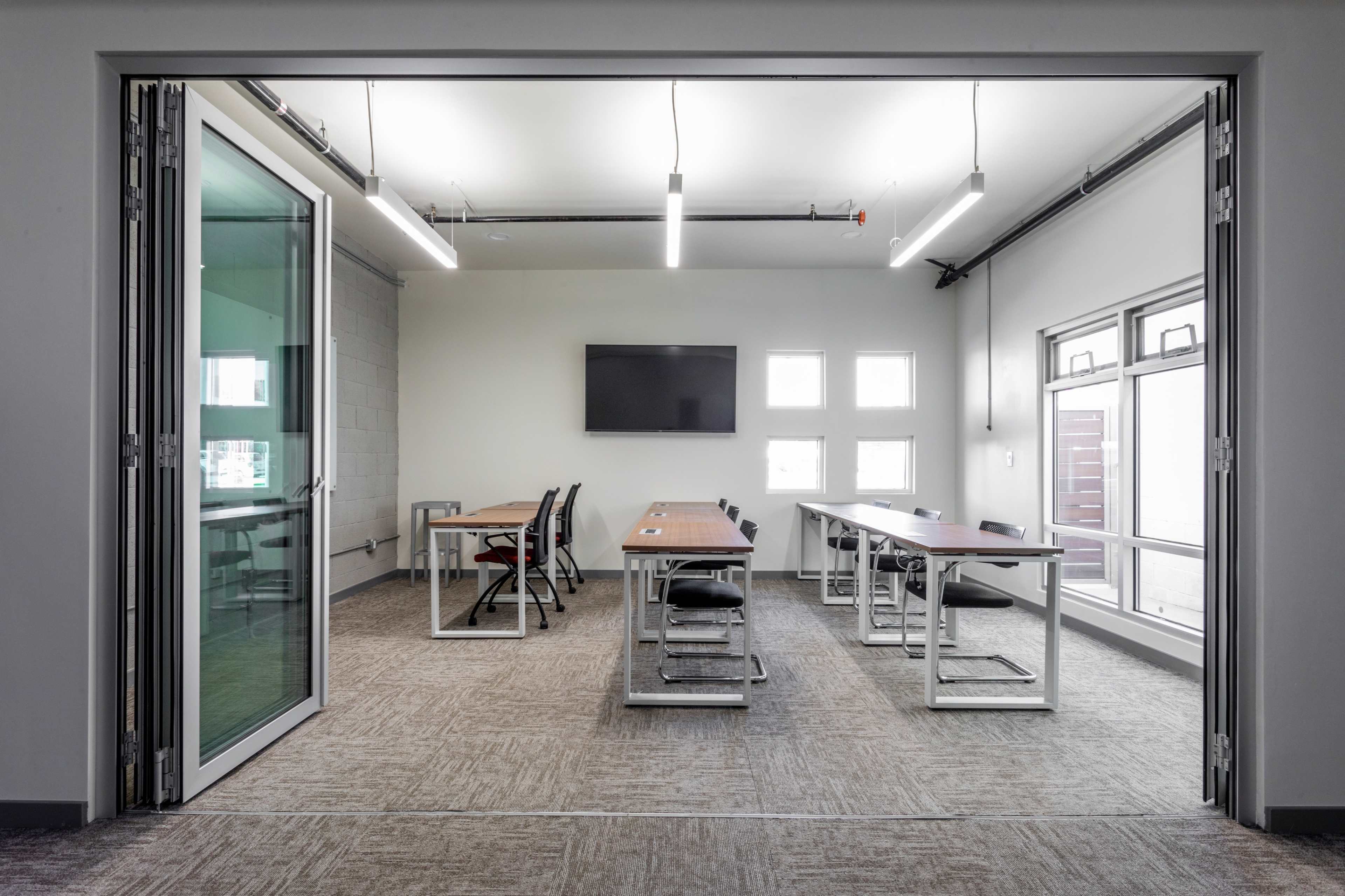 A conference room features four long tables with chairs, a wall-mounted television, and large windows allowing for natural light.