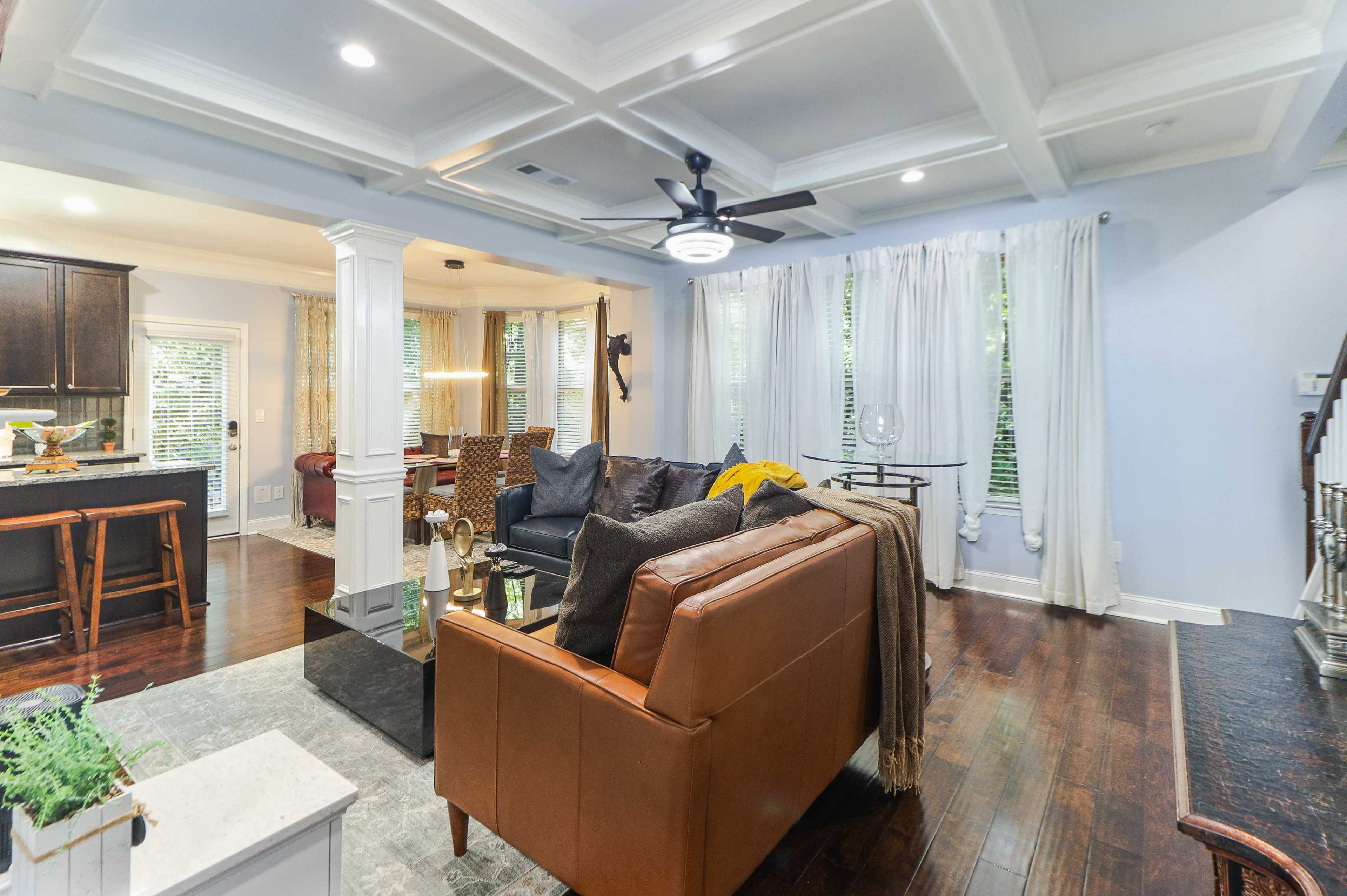 A spacious living room with a brown leather sofa, a coffee table, and a ceiling fan, adjacent to a kitchen area featuring dark cabinets and a dining space.
