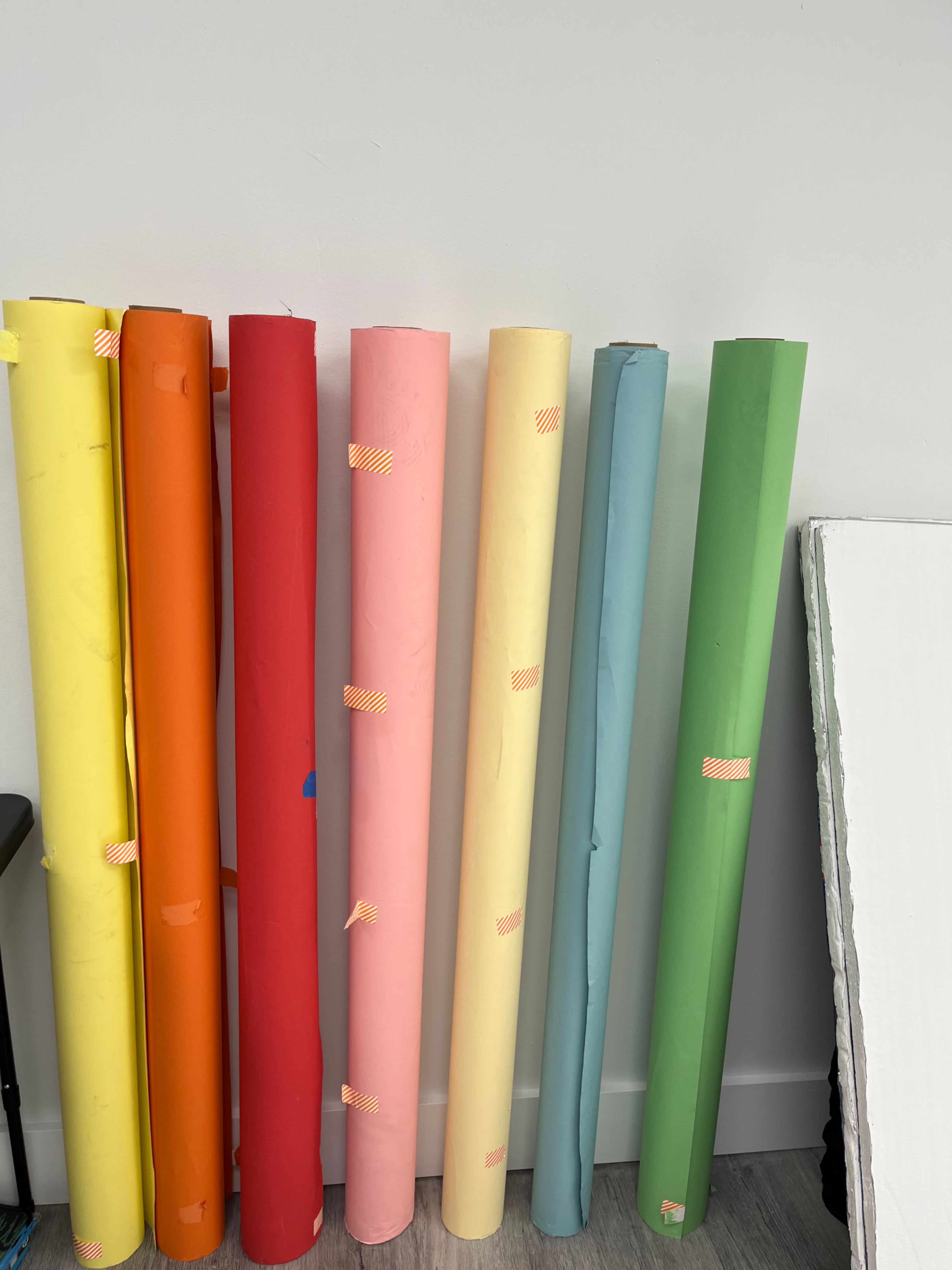 The image shows a row of rolled sheets of colored paper in various hues lined up against a wall.
