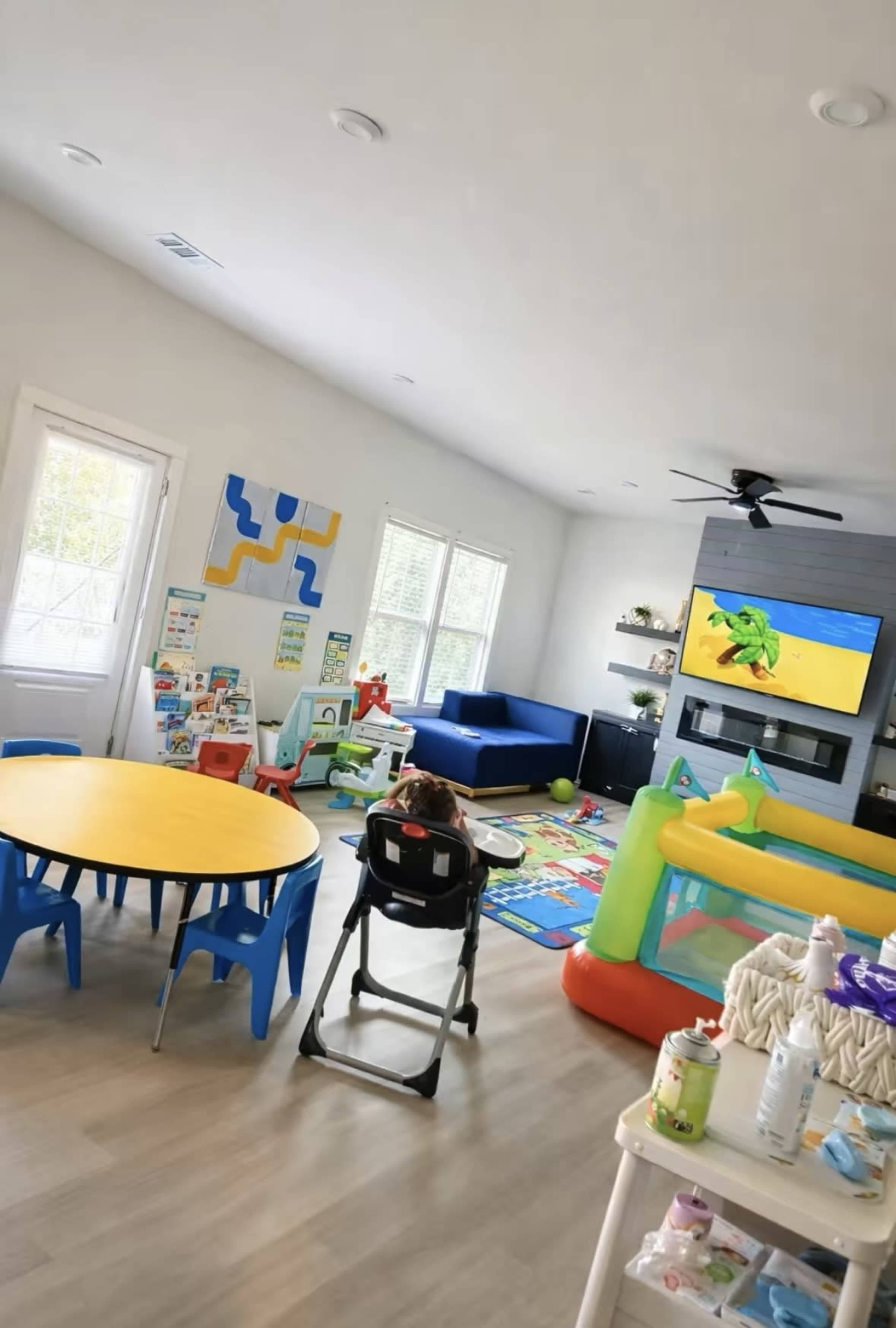 The image shows a bright living room with a high chair, a round yellow table, colorful play mats, and a television displaying a cartoon, along with a play area featuring an inflatable structure.