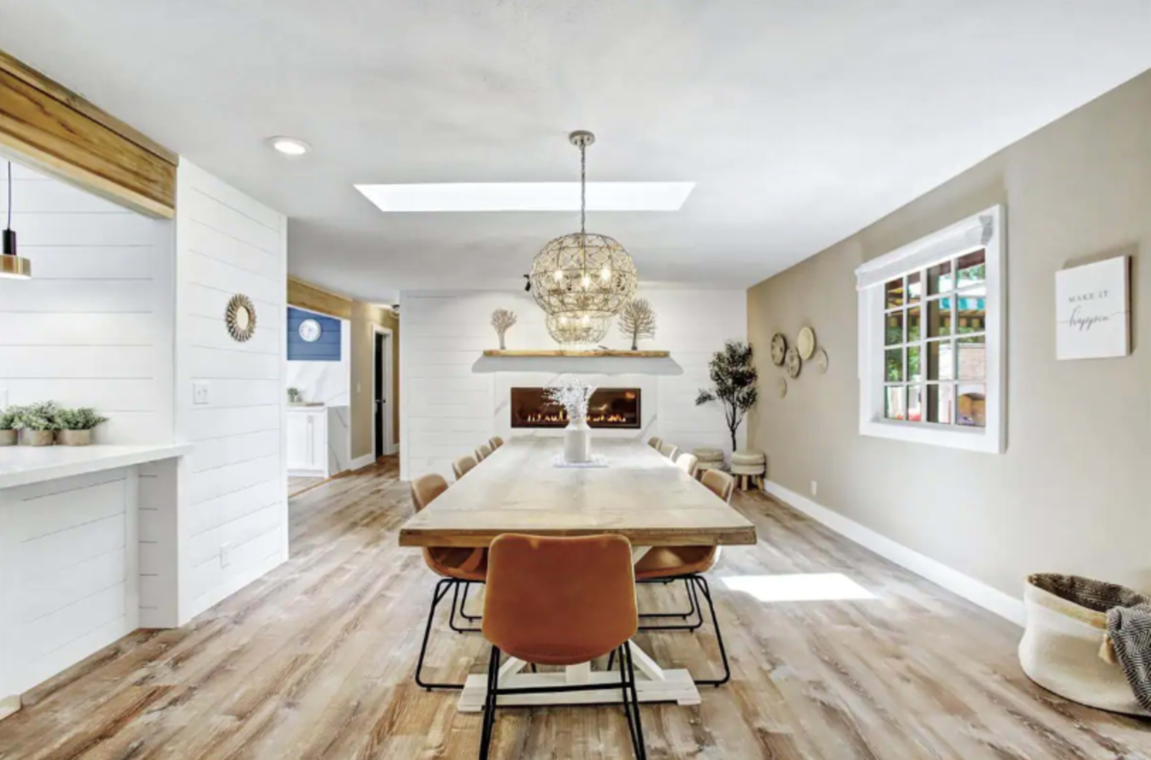 A modern dining room with a long wooden table surrounded by brown chairs, illuminated by a large pendant light, and a cozy fireplace visible in the background.