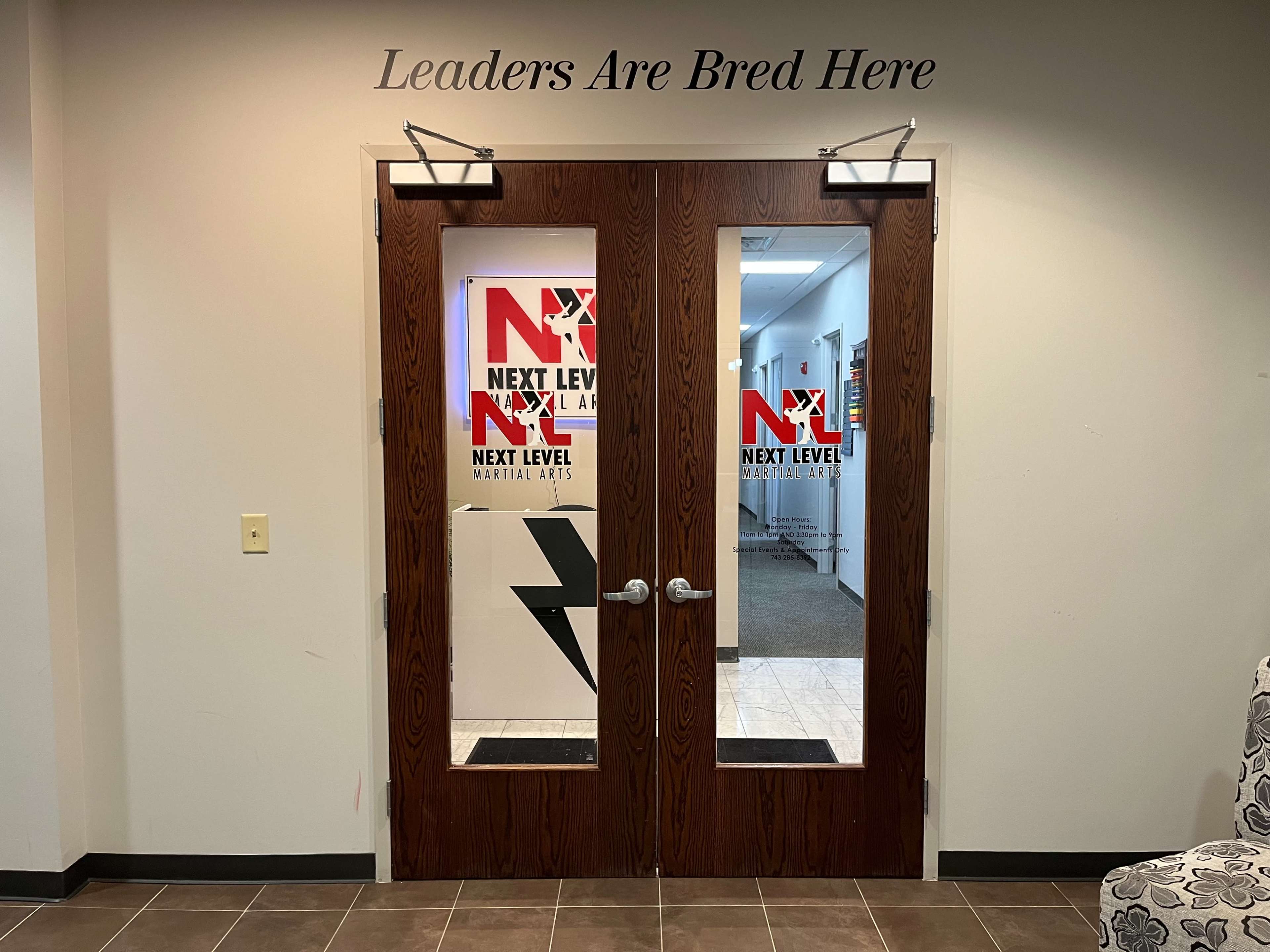 The image shows double doors with glass panels that display the logos of a martial arts studio, framed by a wall with the phrase "Leaders Are Bred Here" above them.