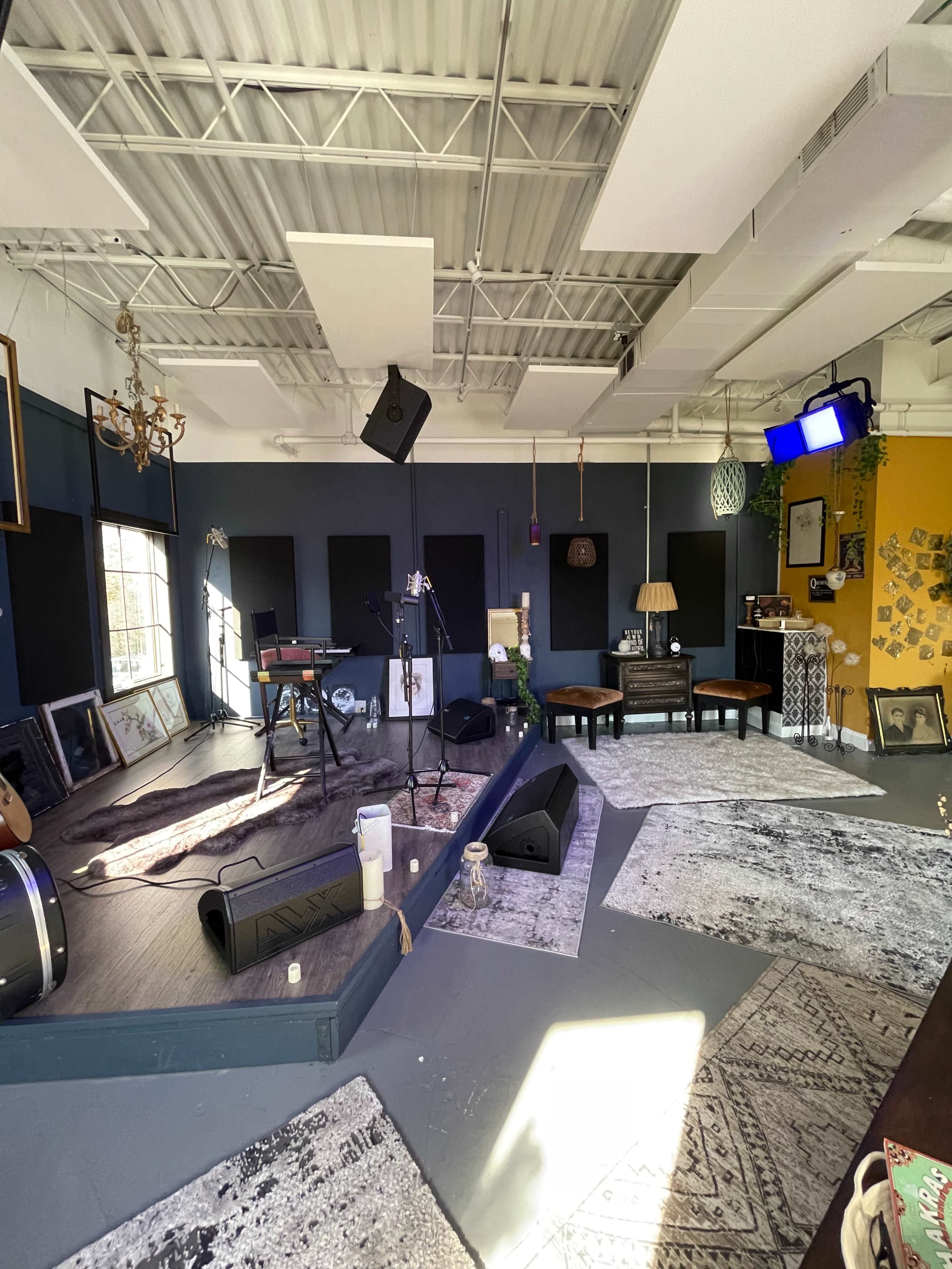 A music studio with a stage area featuring instruments, microphones, and various furniture arranged around a wooden floor.