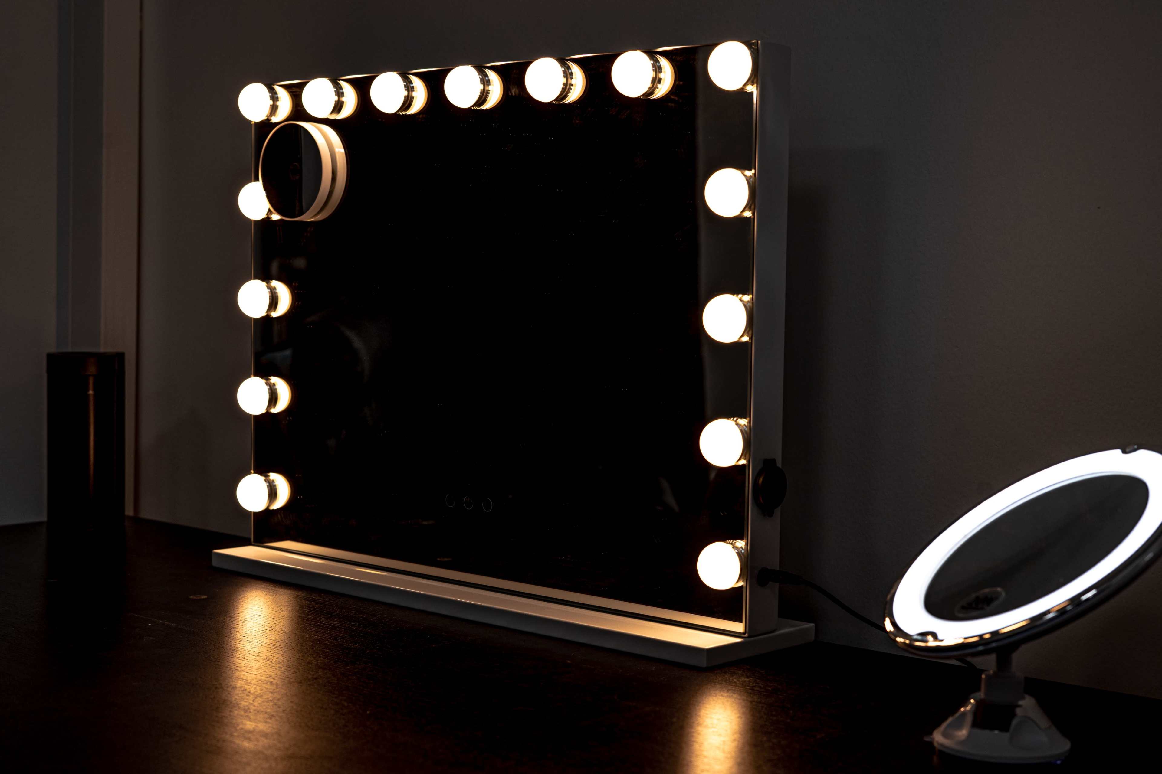 A rectangular makeup mirror with bright circular bulbs surrounds its frame is positioned on a dark surface next to a smaller illuminated mirror.