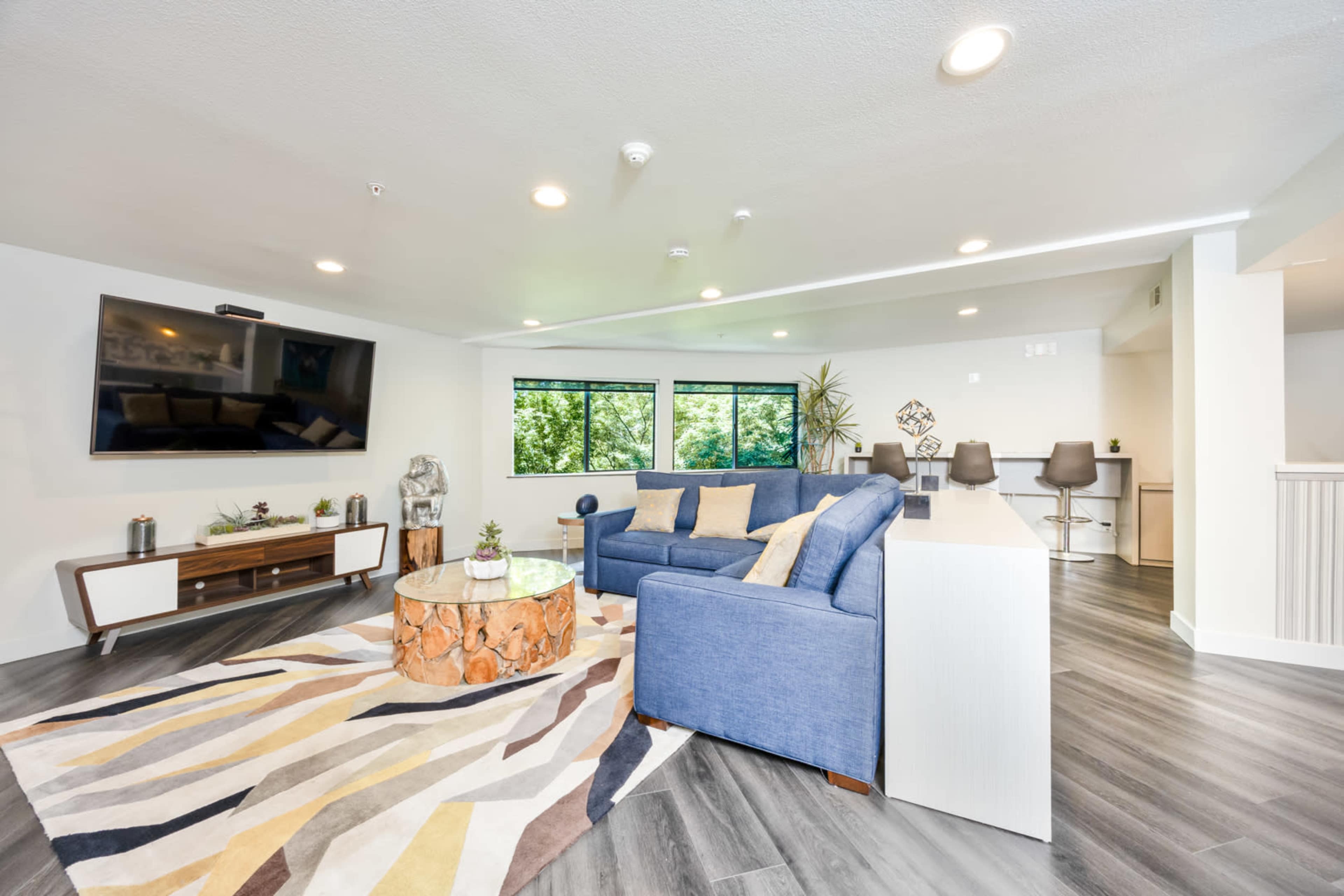 A modern living room features a blue sofa, a circular wooden coffee table, a large television mounted on the wall, and a rug with geometric patterns, all set against windows that overlook greenery.