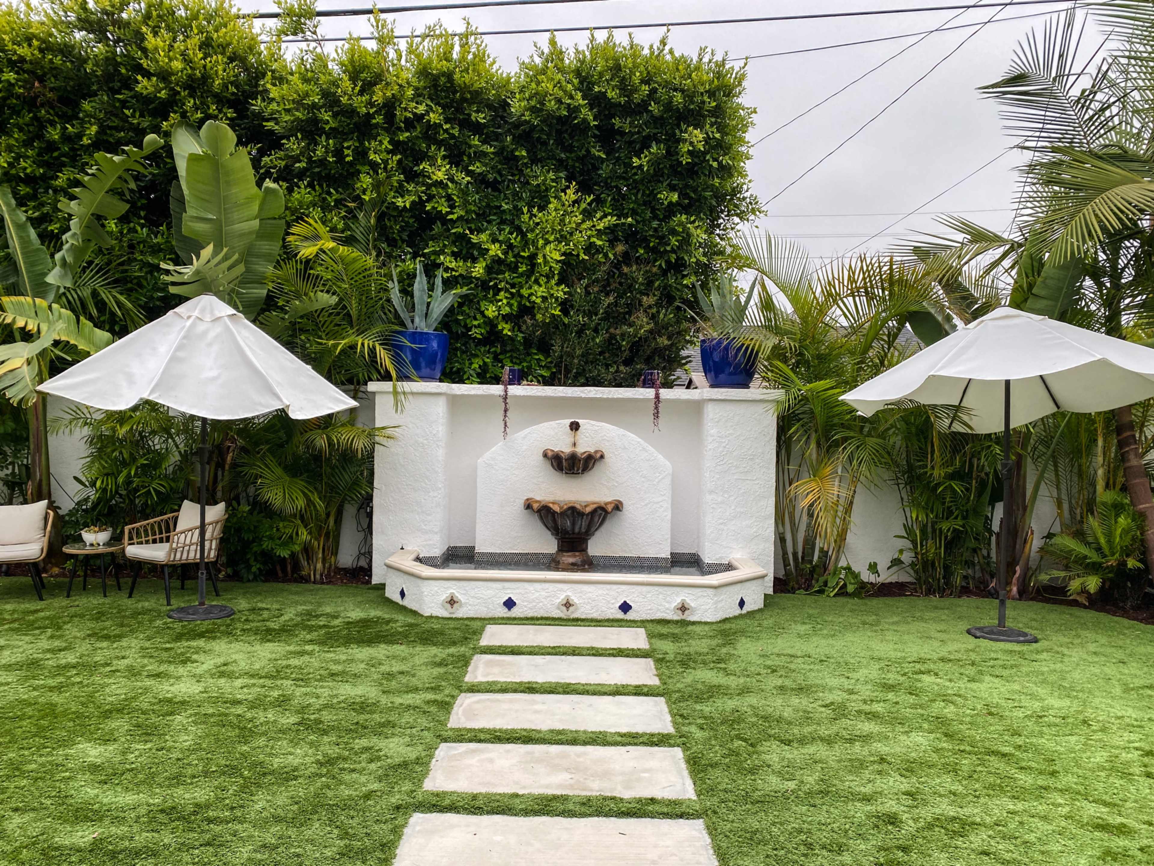 A landscaped garden features a tiered fountain, surrounded by tropical plants and two white umbrellas over seating areas.