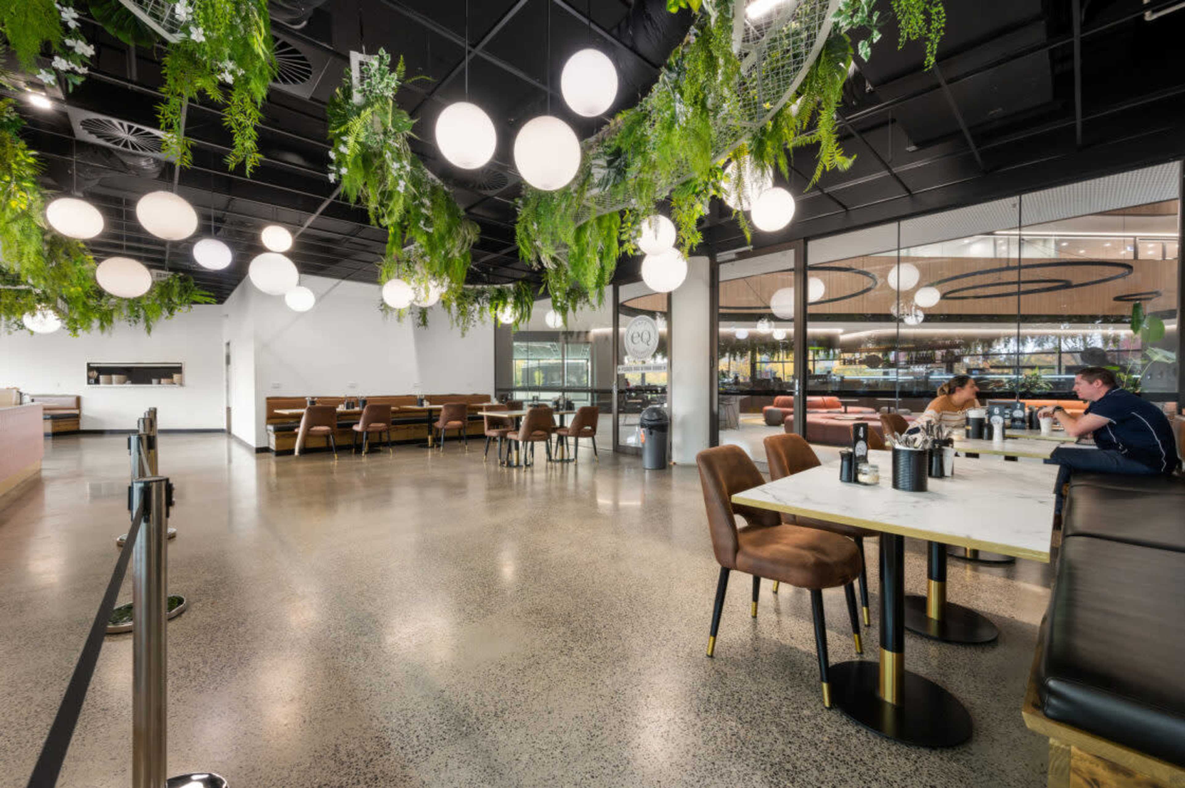 The image shows a modern cafe with hanging greenery, round light fixtures, and a mix of seating arrangements, including tables and benches.