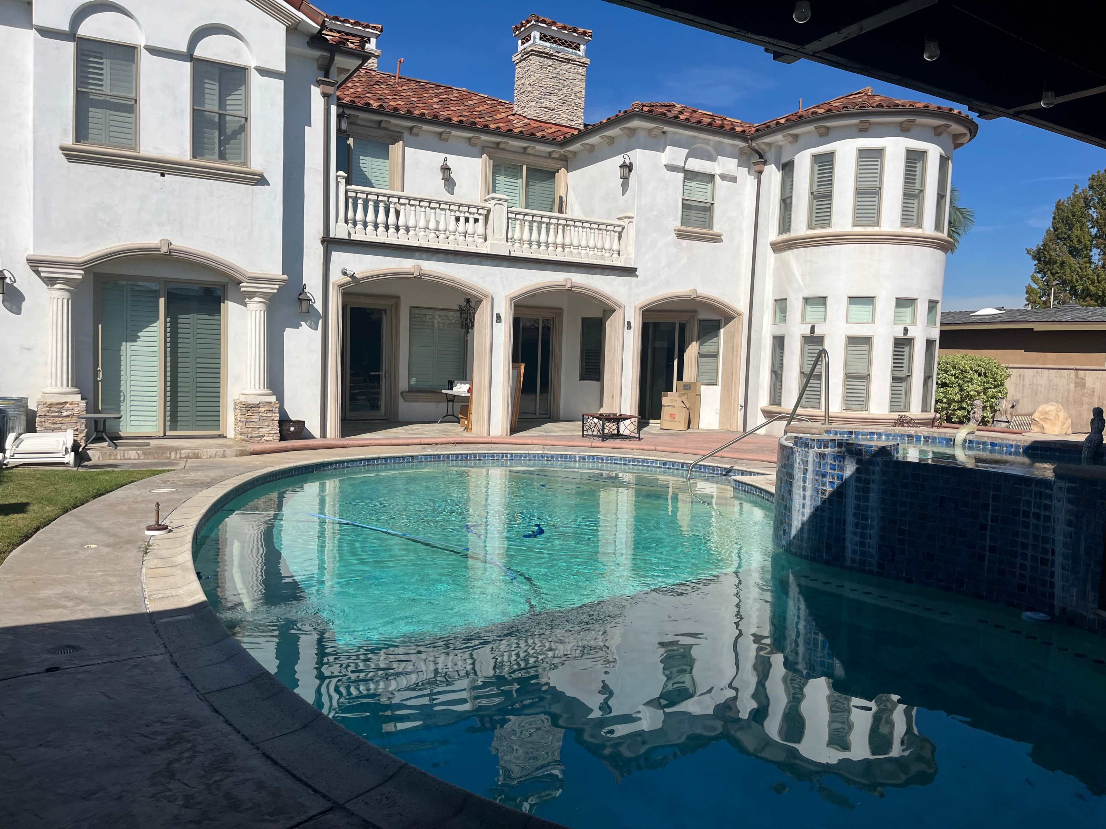 A clear swimming pool reflects the exterior of a large, two-story house with a stucco facade and a tiled roof.
