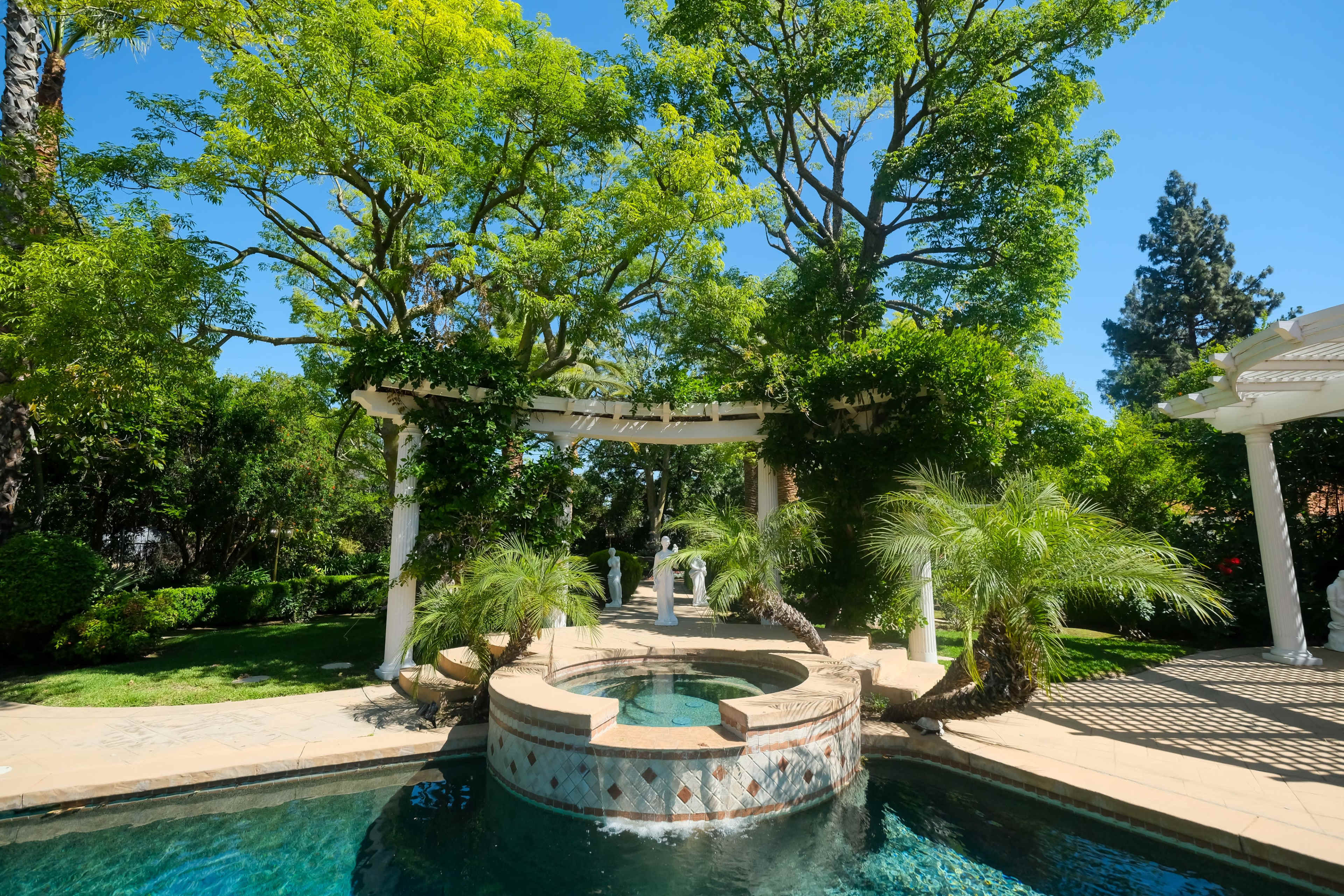 A landscaped garden features a circular stone pool surrounded by lush greenery, large trees, and a pergola adorned with vines.