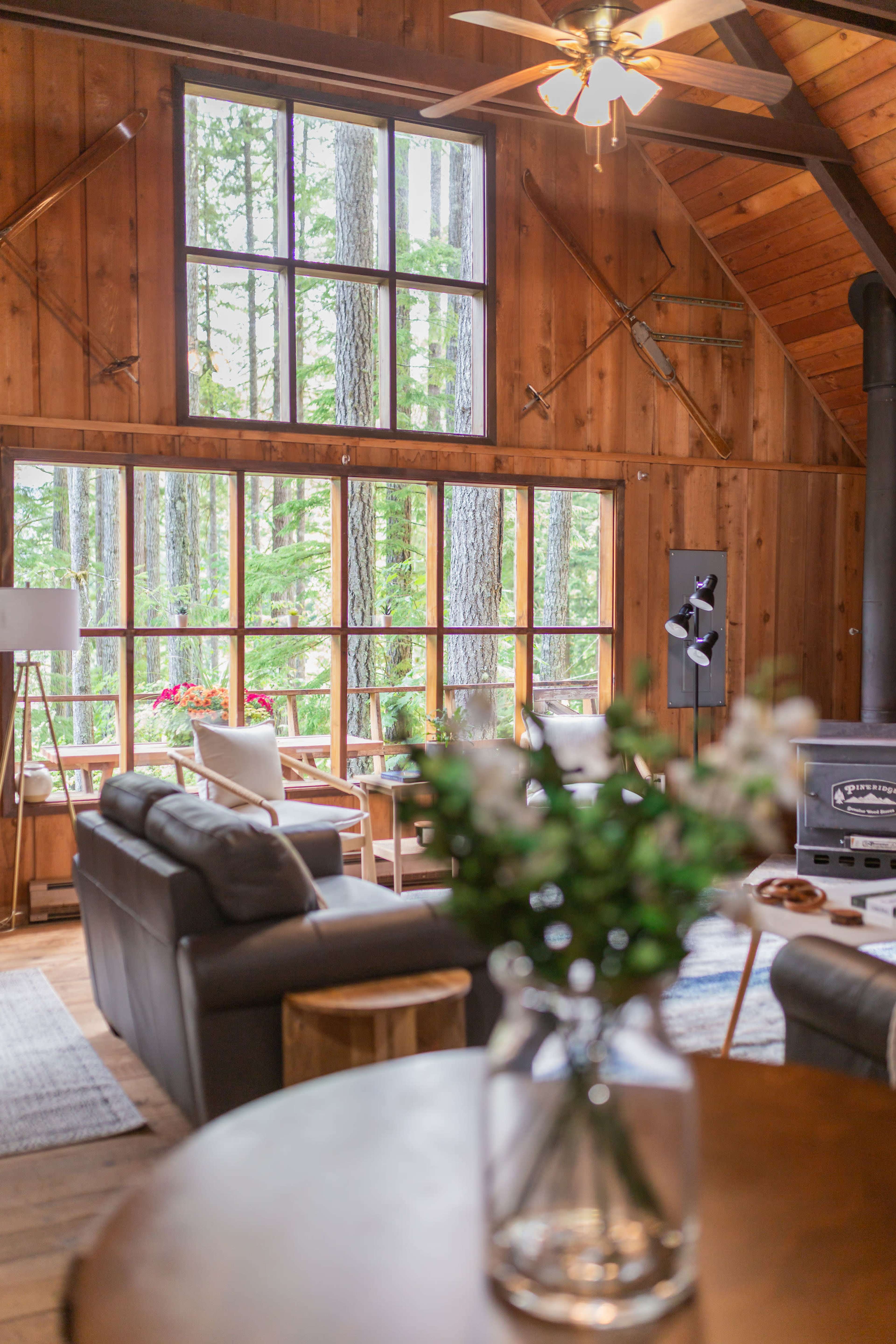 A cozy wooden cabin interior featuring a living area with a leather sofa, a coffee table, and large windows that overlook a forested view.