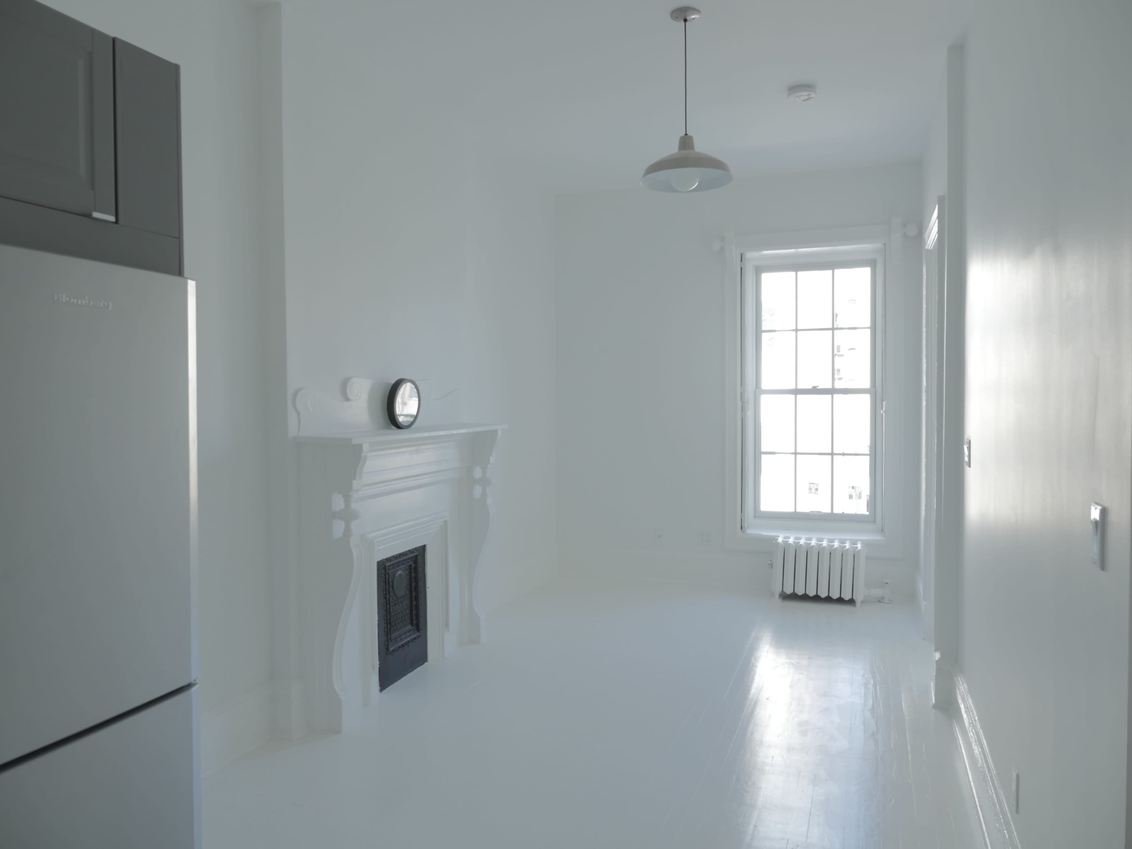 A bright, minimally furnished room features a white fireplace, a large window, and a stainless steel refrigerator.