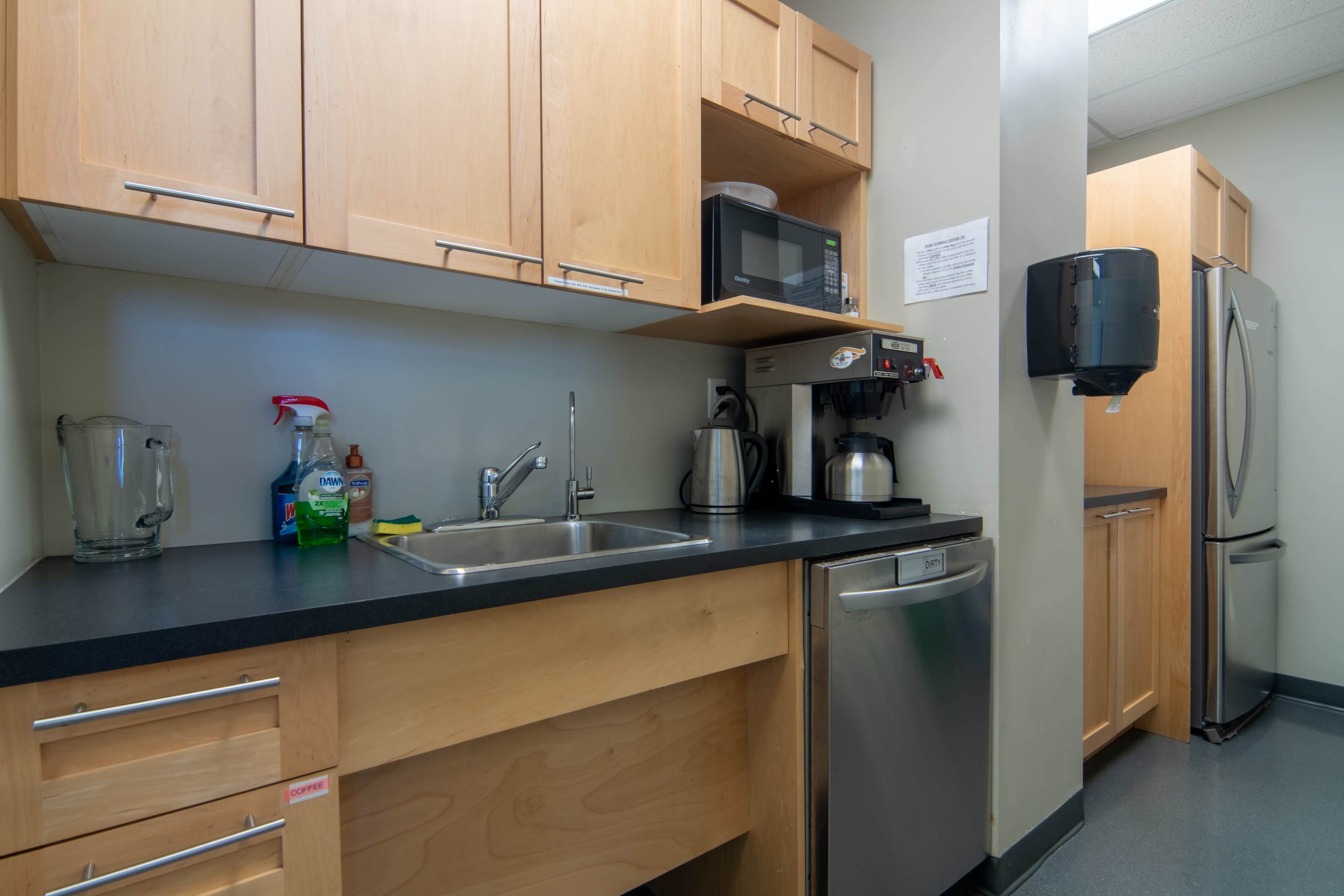 The image shows a kitchenette featuring wooden cabinets, a sink, a microwave, a coffee maker, and a stainless steel refrigerator.