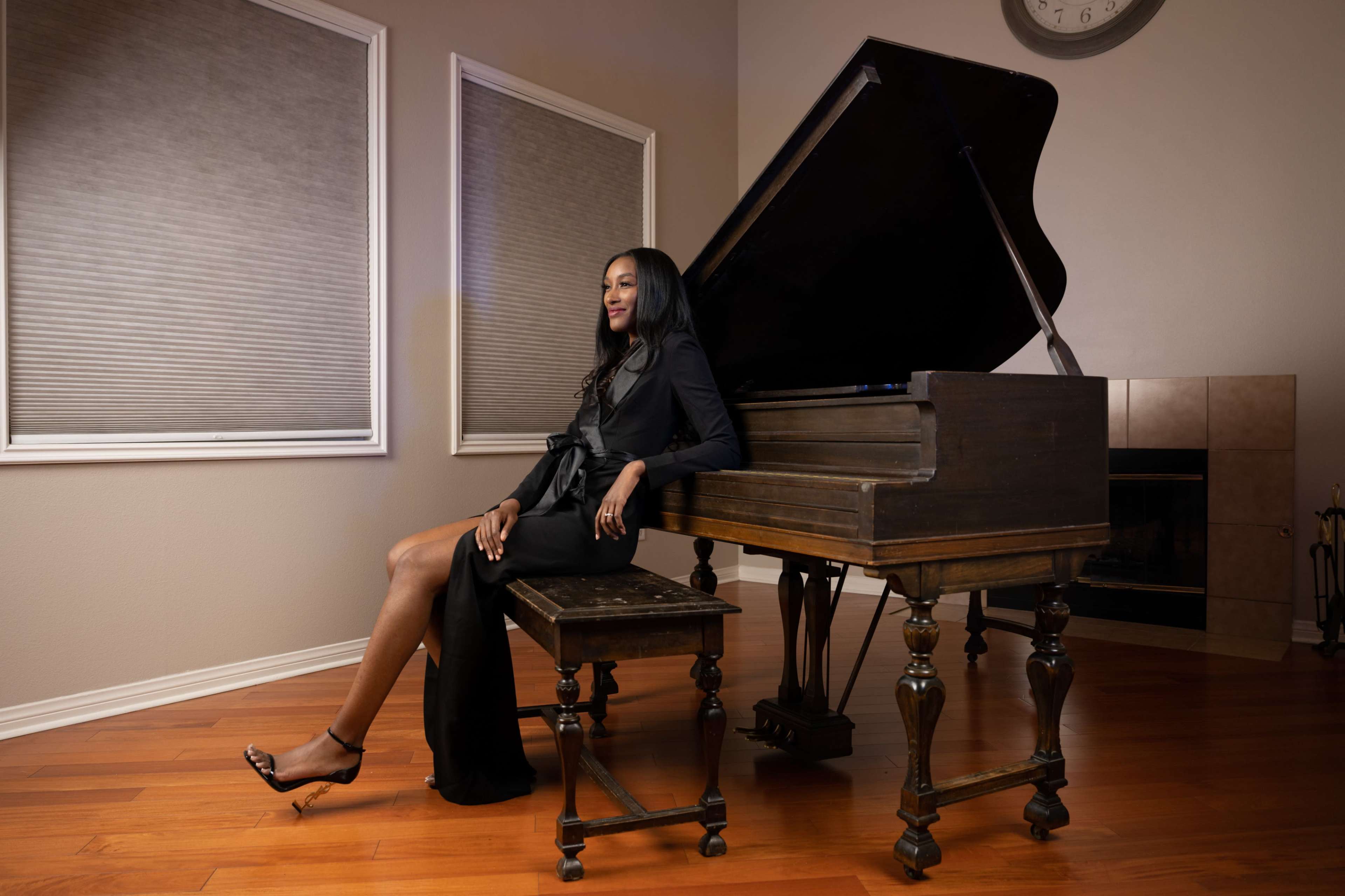 A woman in a black dress sits elegantly beside a grand piano in a well-lit room with wooden flooring.