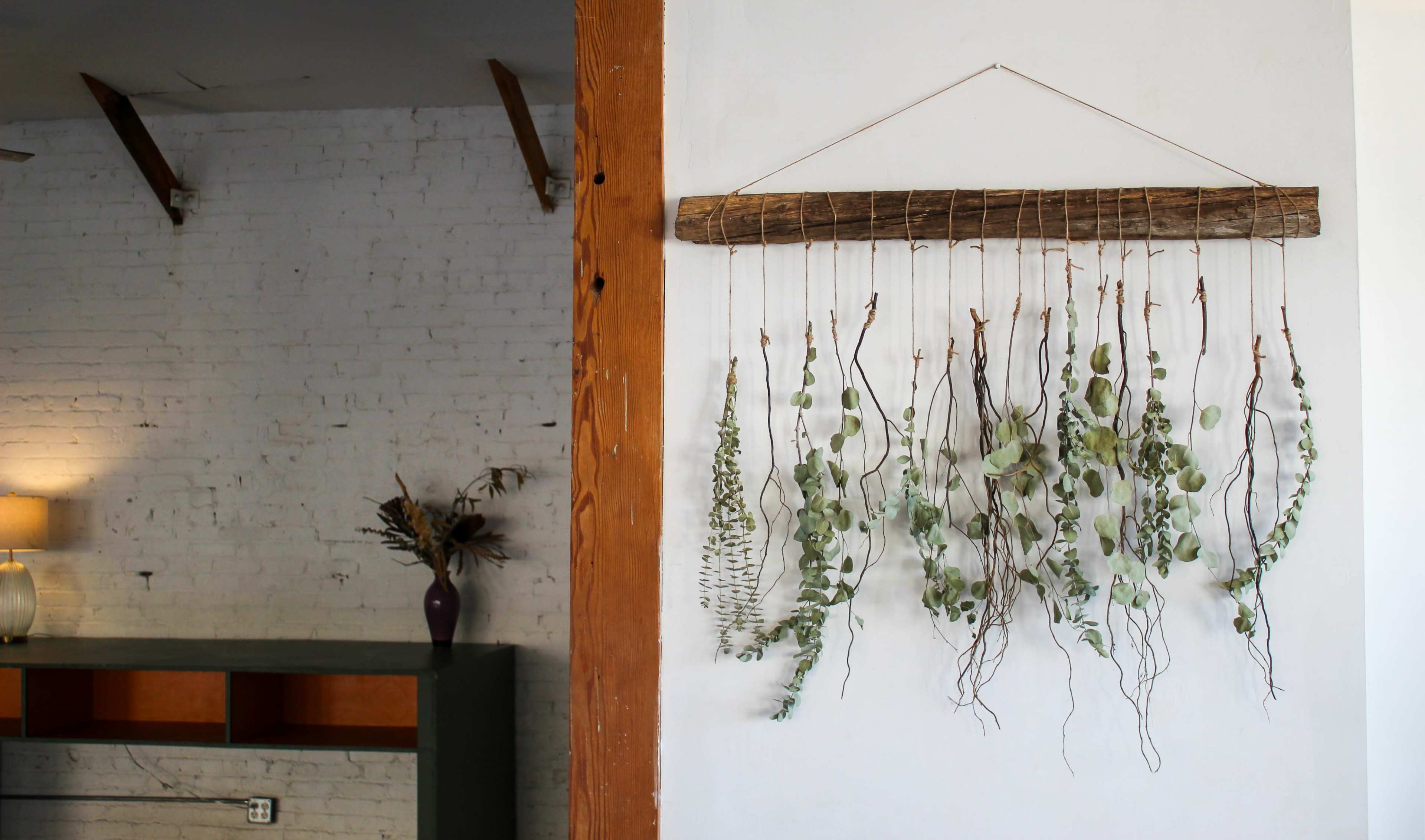 The image shows a decorative wall hanging made of dried greenery suspended from a wooden rod, with a simple interior setting in the background.