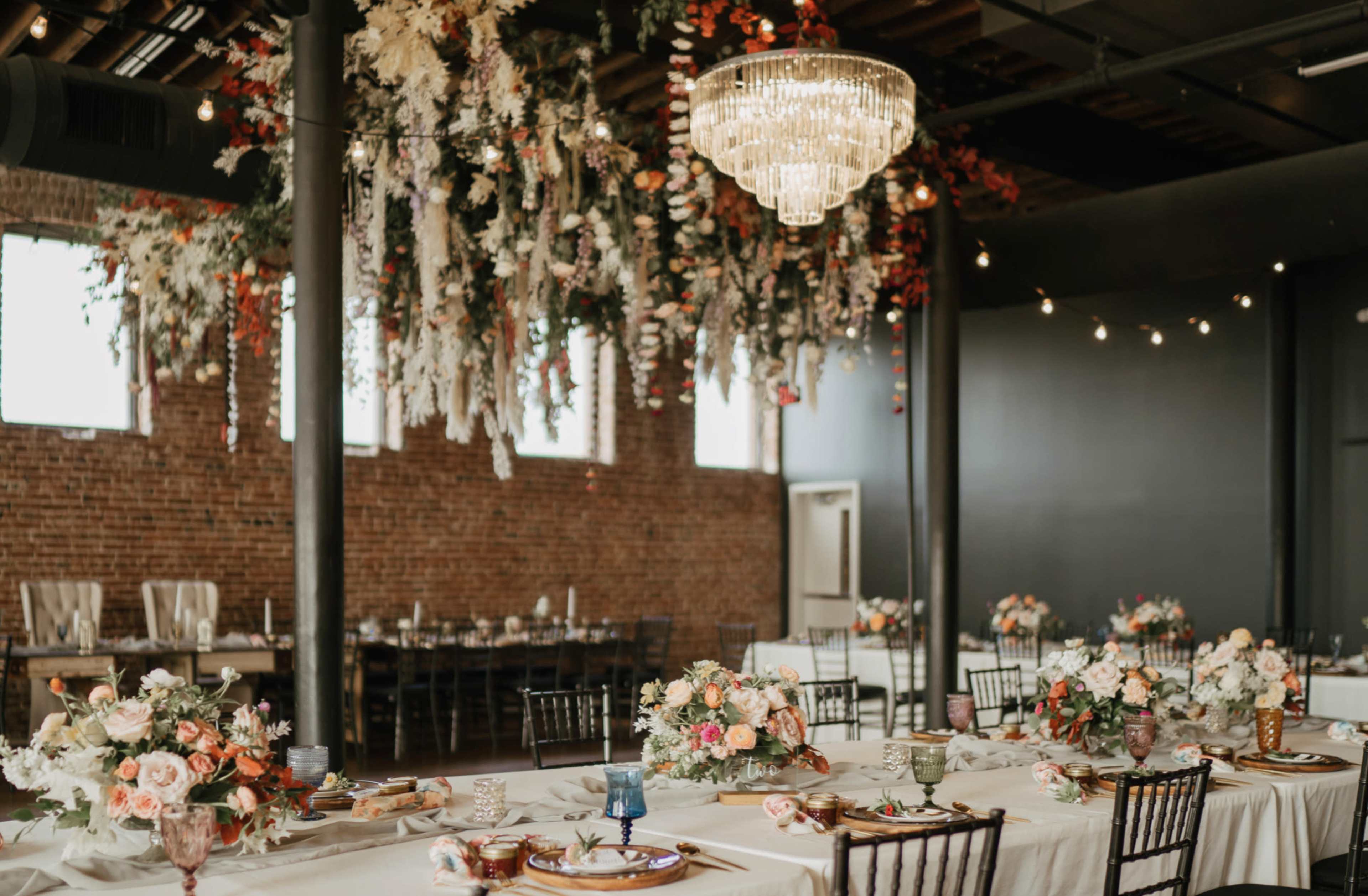 The image shows a decorated dining venue with tables set for a formal event, featuring floral centerpieces and a large chandelier suspended from the ceiling.