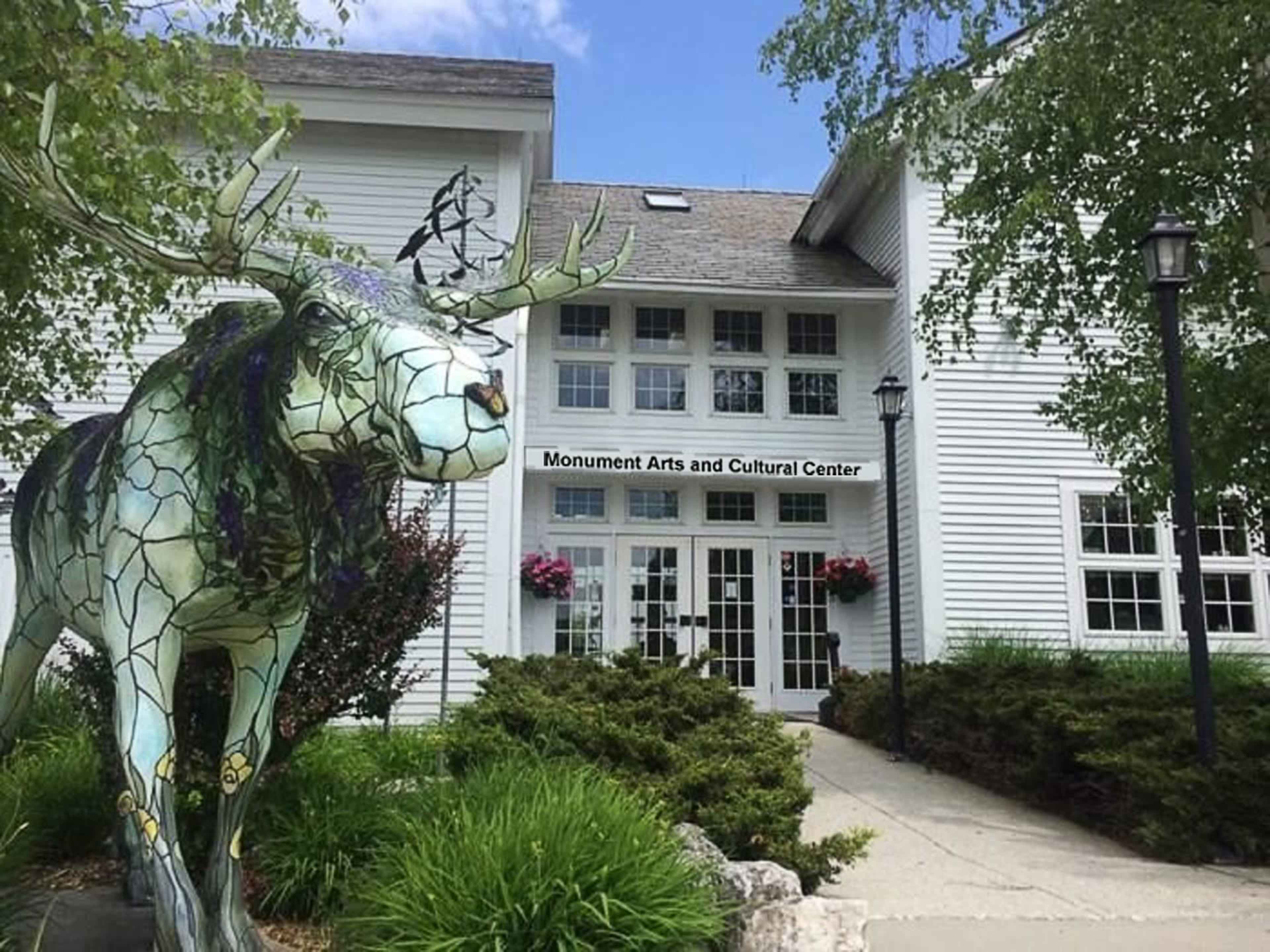 A colorful, mosaic-style moose sculpture in front of the Monument Arts and Cultural Center, a white building with large windows and flower baskets.