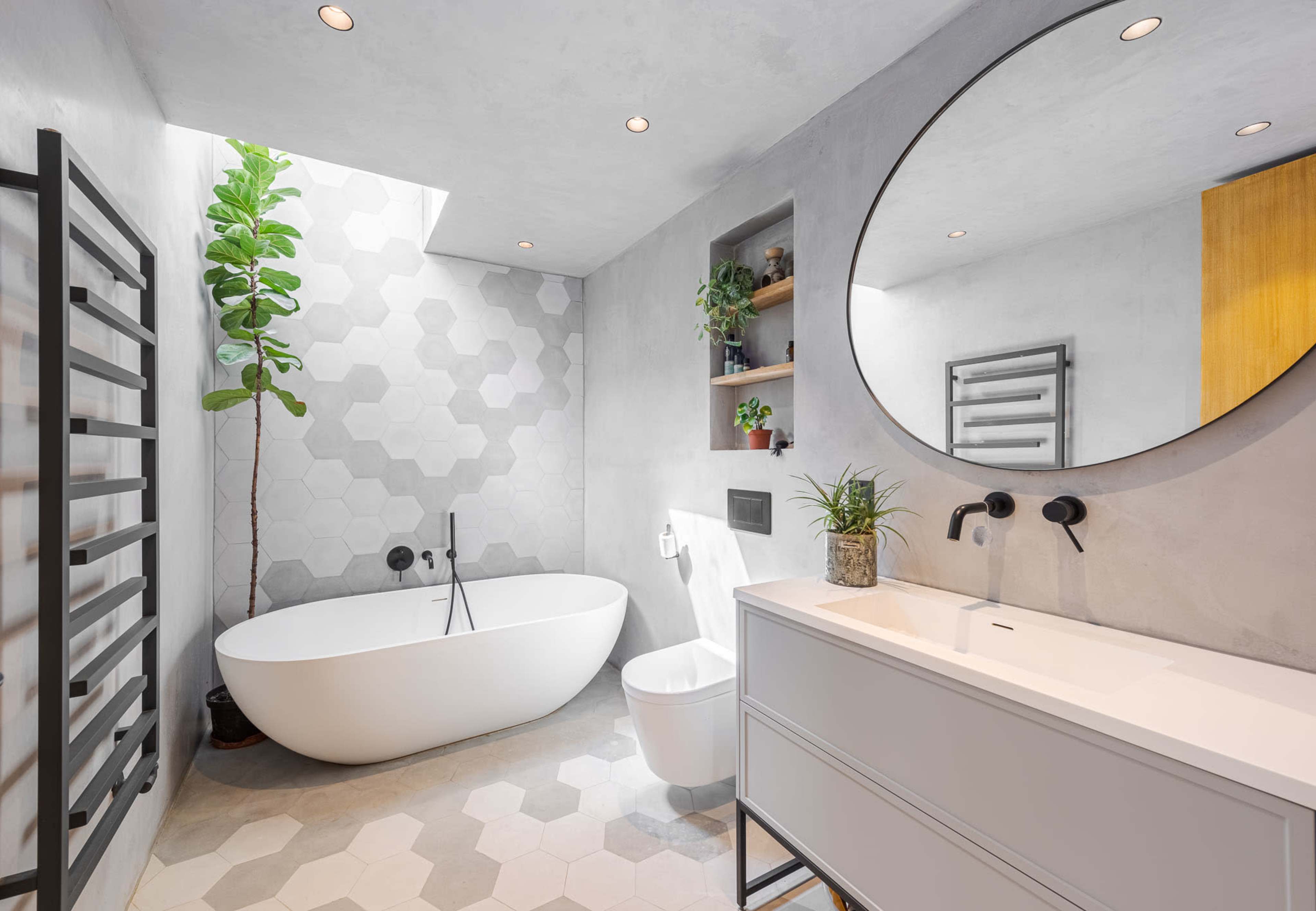 The bathroom features a freestanding bathtub, a circular mirror above a vanity, and hexagonal tile flooring with a plant positioned near a skylight.