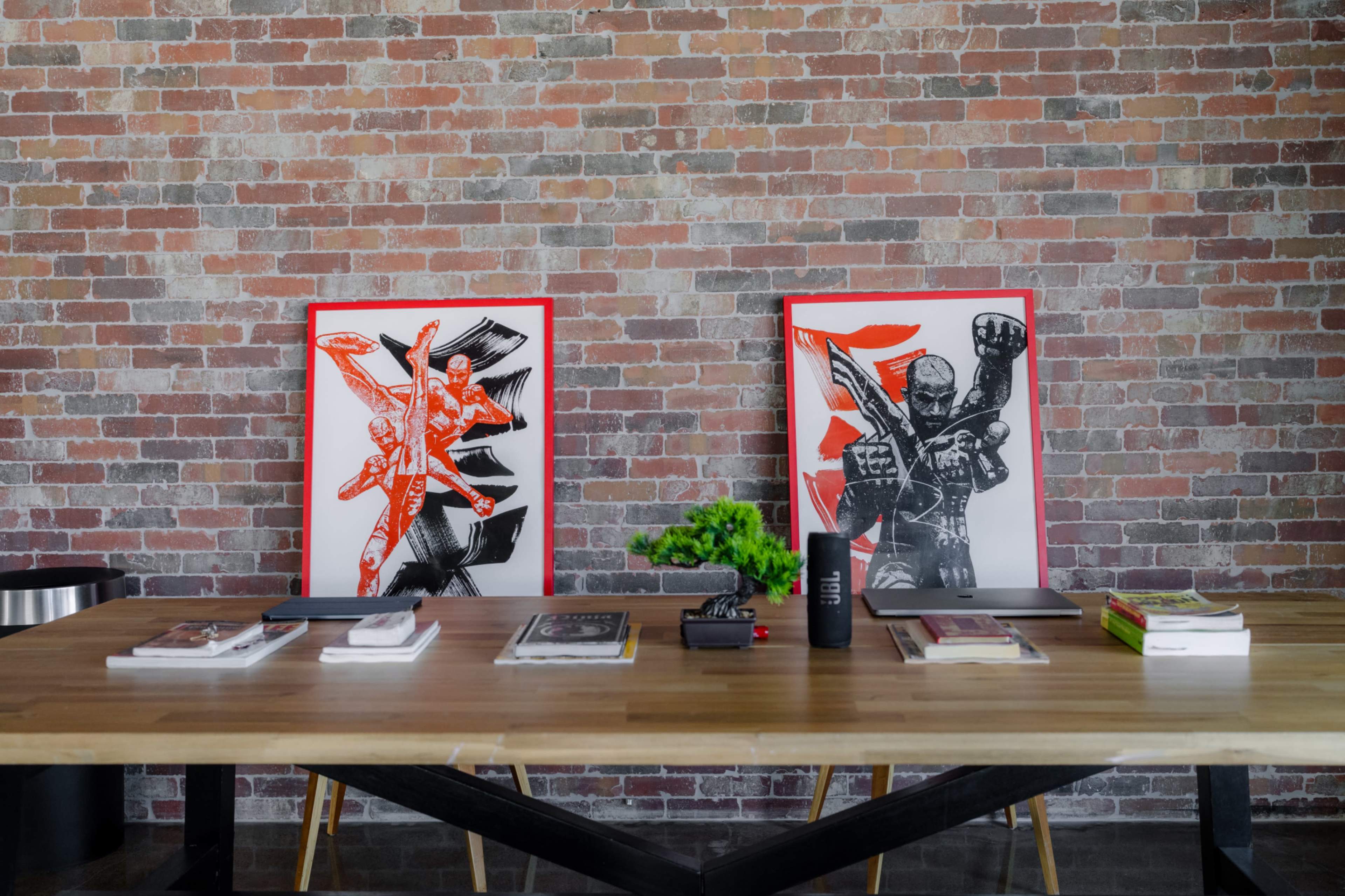 The image features a wooden table displaying various books and magazines, with two bold graphic art pieces on the wall behind it, set against a backdrop of exposed brick.
