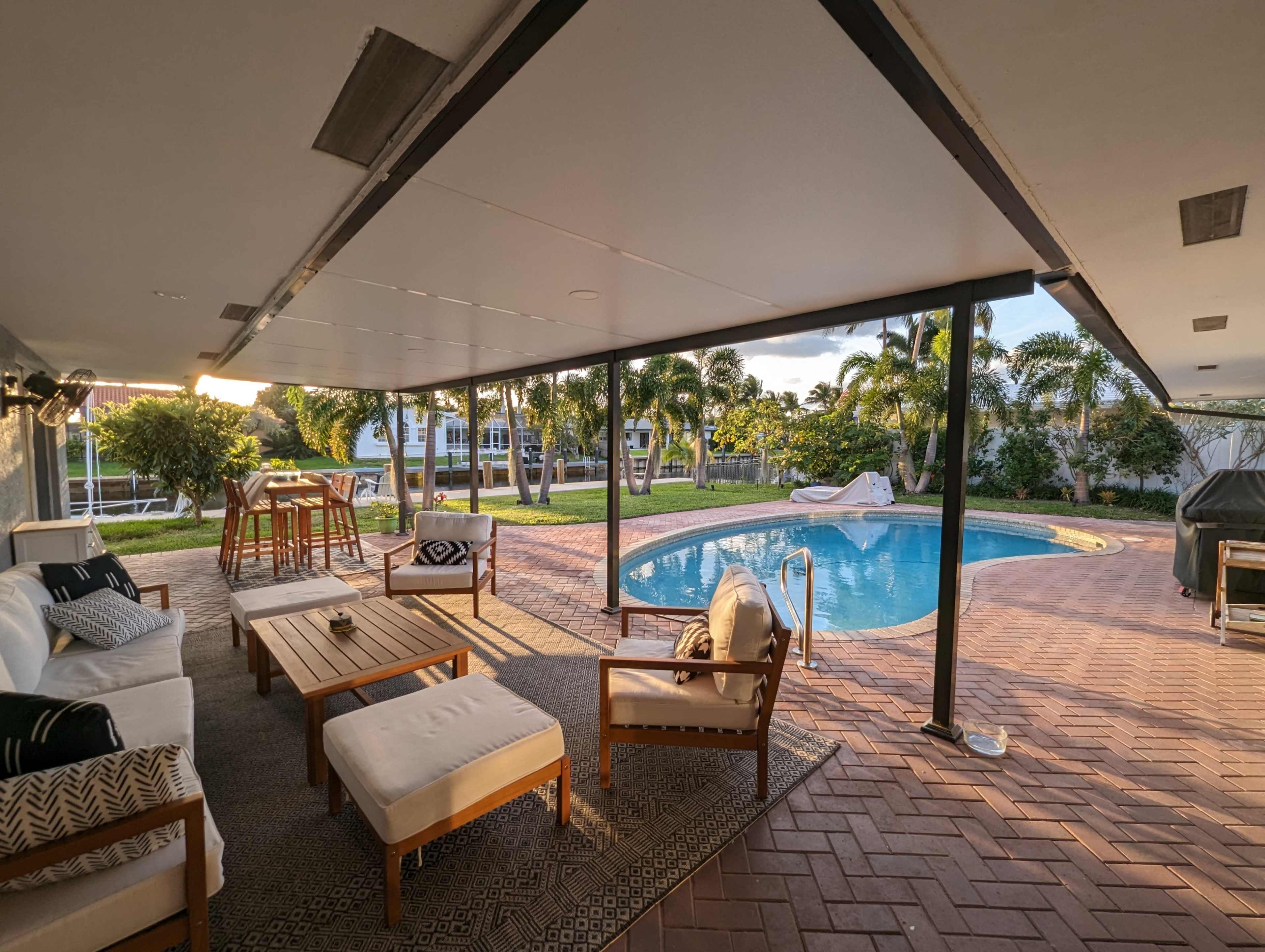 A covered patio with wooden furniture overlooks a swimming pool surrounded by palm trees and a well-maintained lawn.