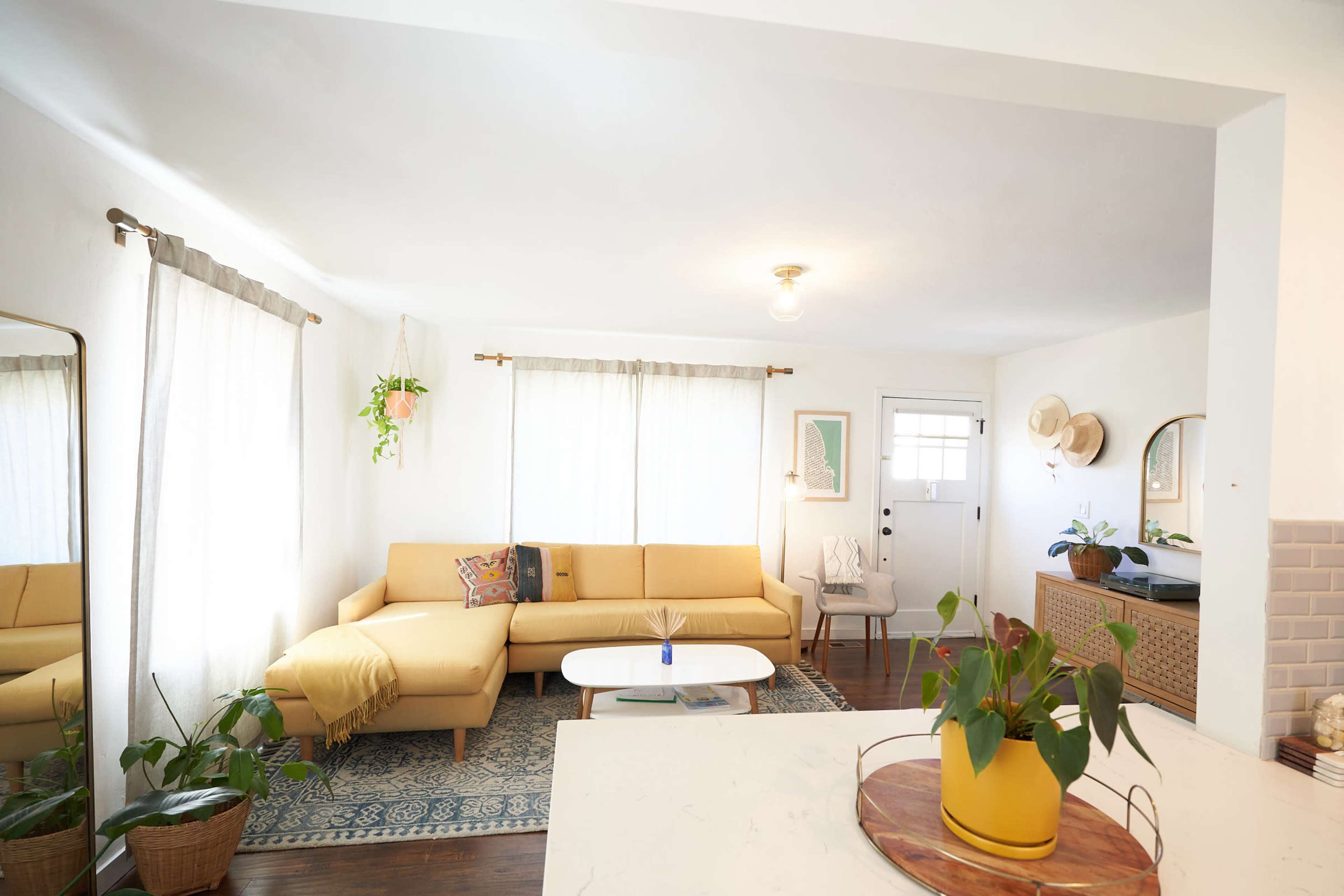 A cozy living room features a yellow sectional sofa, a light wooden coffee table, and large windows covered with sheer curtains.