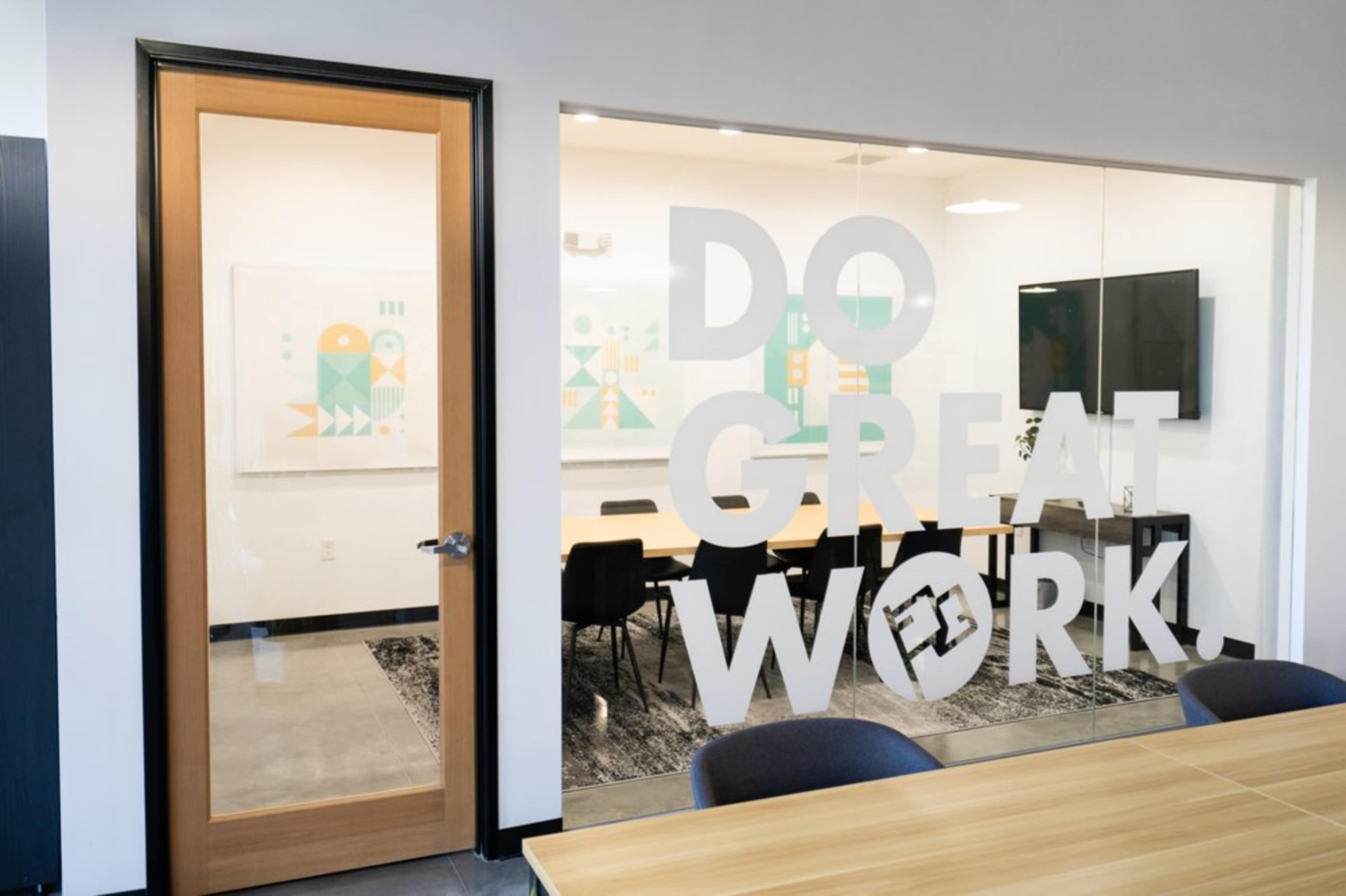 A modern conference room with a large glass wall displaying the phrase "DO GREAT WORK!" and a view of a long table surrounded by chairs.