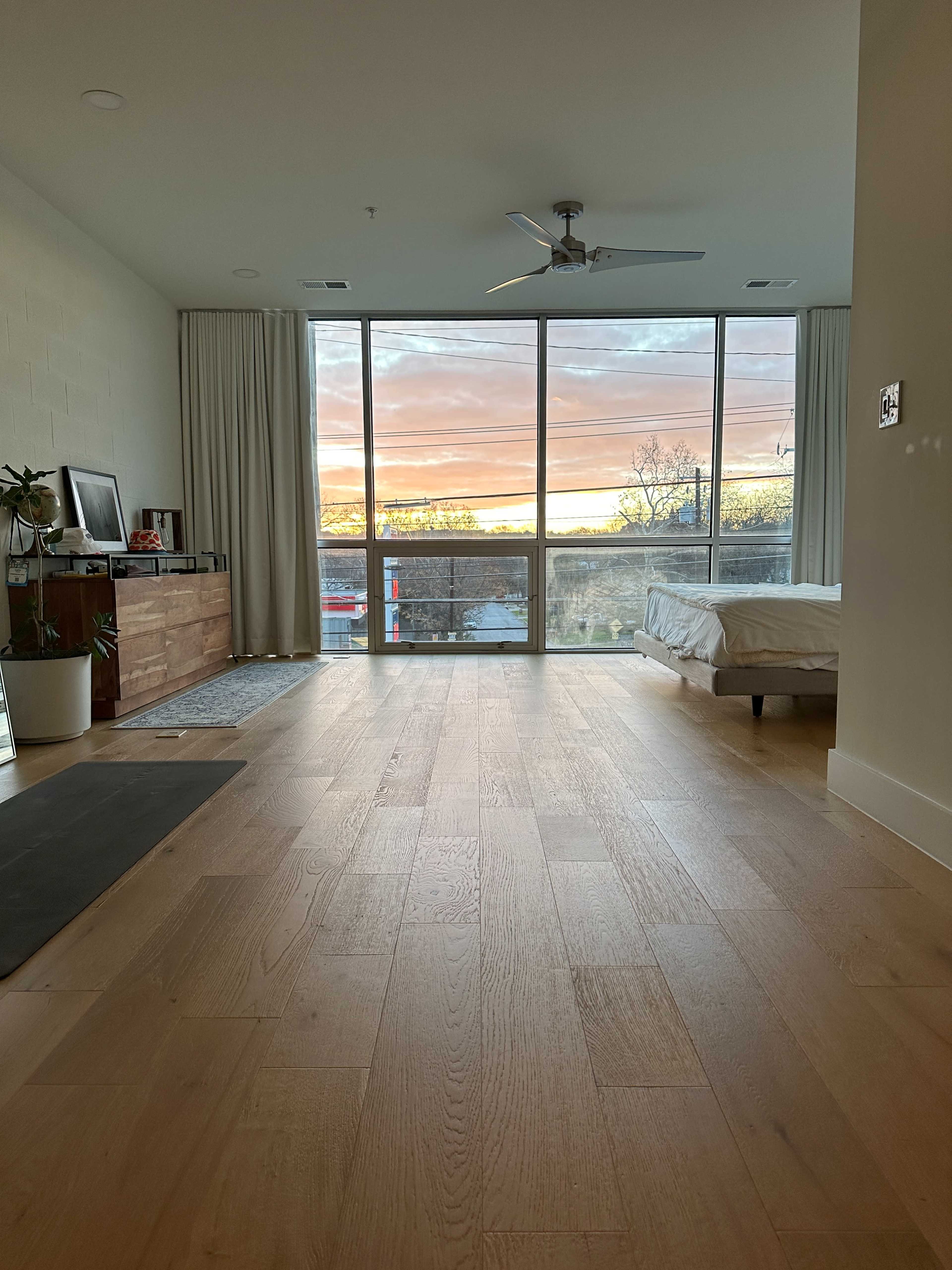 A minimalist bedroom features a large window with a view of a colorful sunset and light wooden flooring.