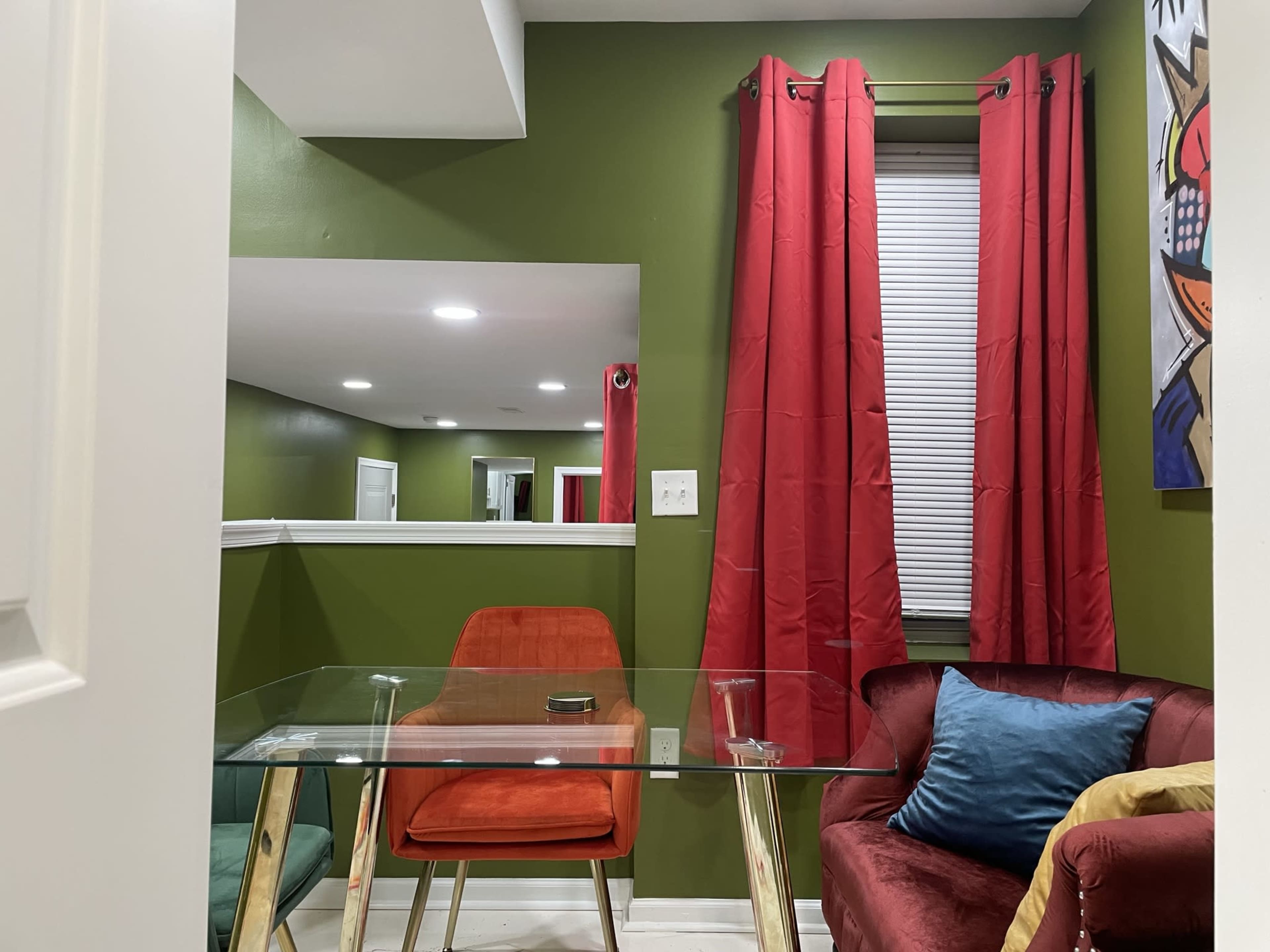 A small room with a glass table, an orange chair, and a red couch against green walls, with red curtains framing a window.