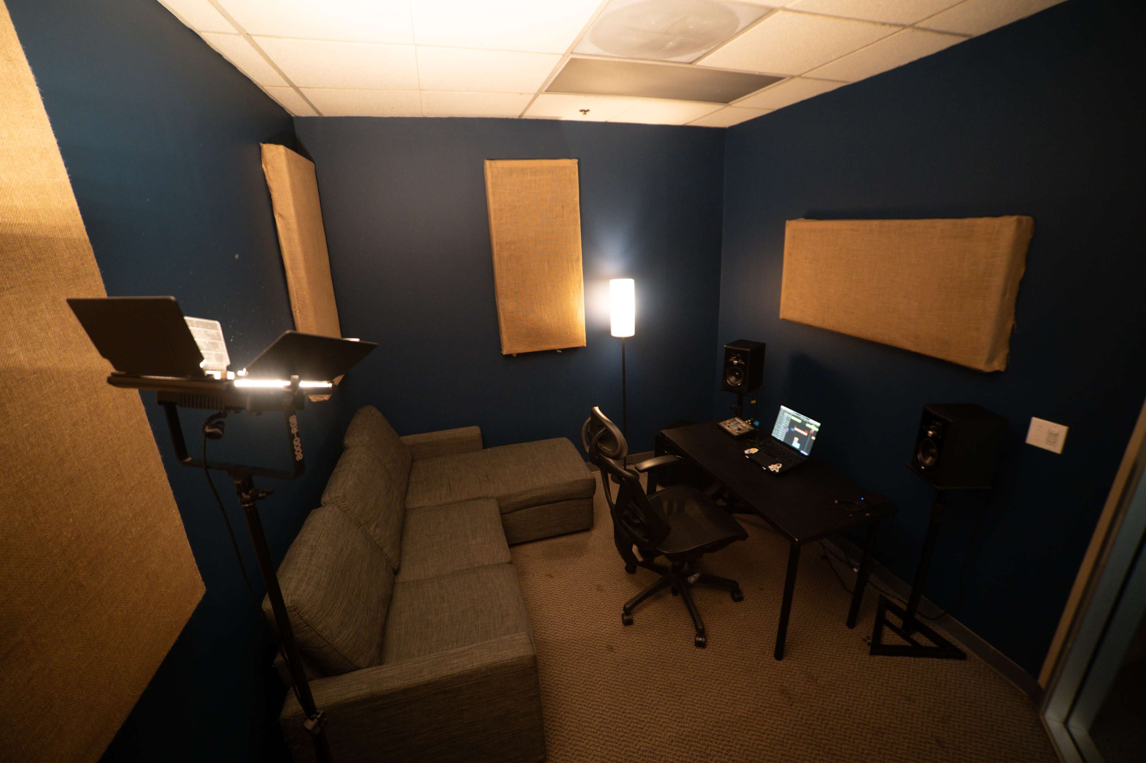 The image shows a small recording studio with a couch, a desk with speakers, and soundproofing panels on the walls.