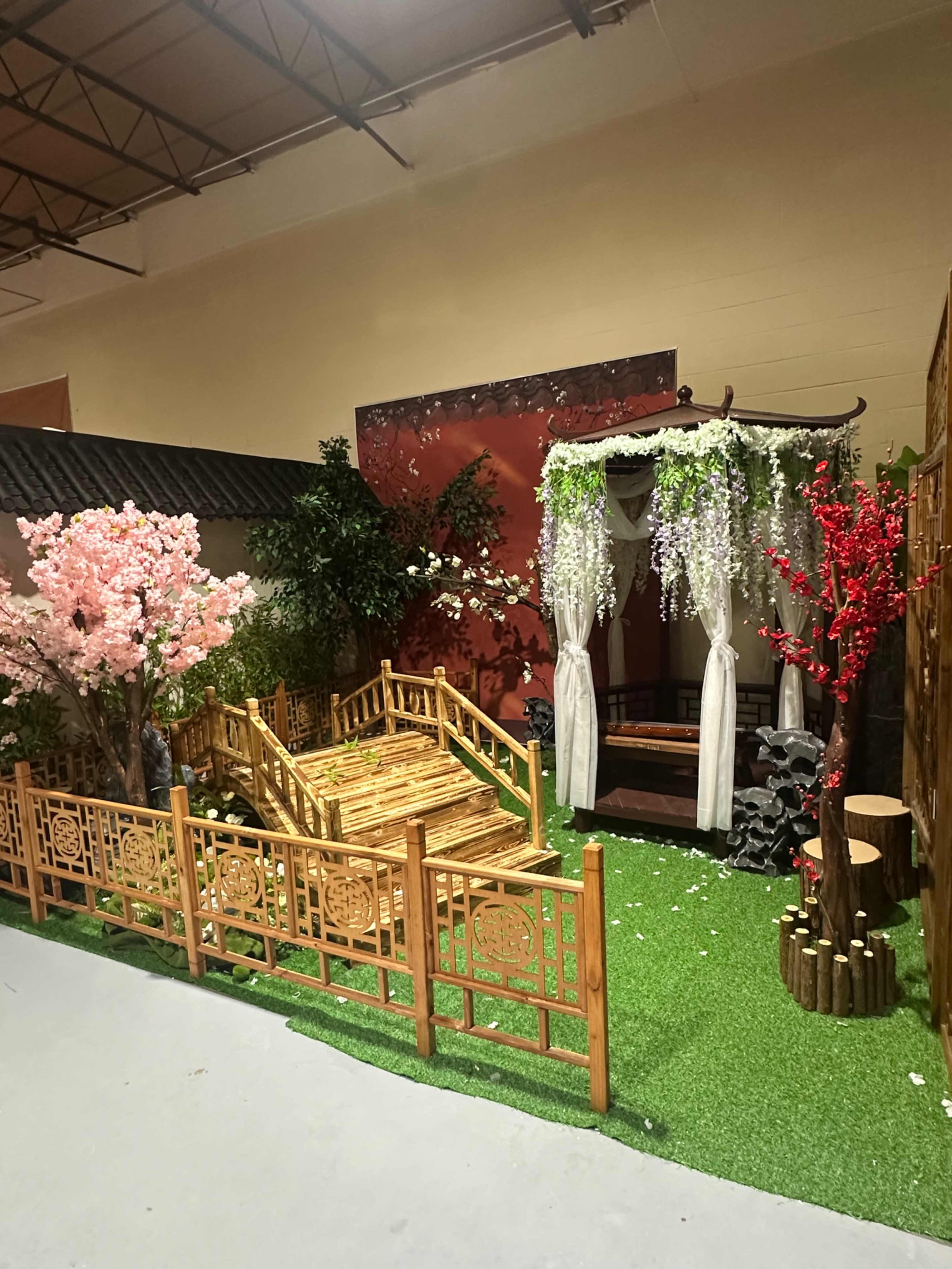The image shows a decorative garden scene featuring a wooden gazebo surrounded by floral trees, greenery, and a bamboo fence.