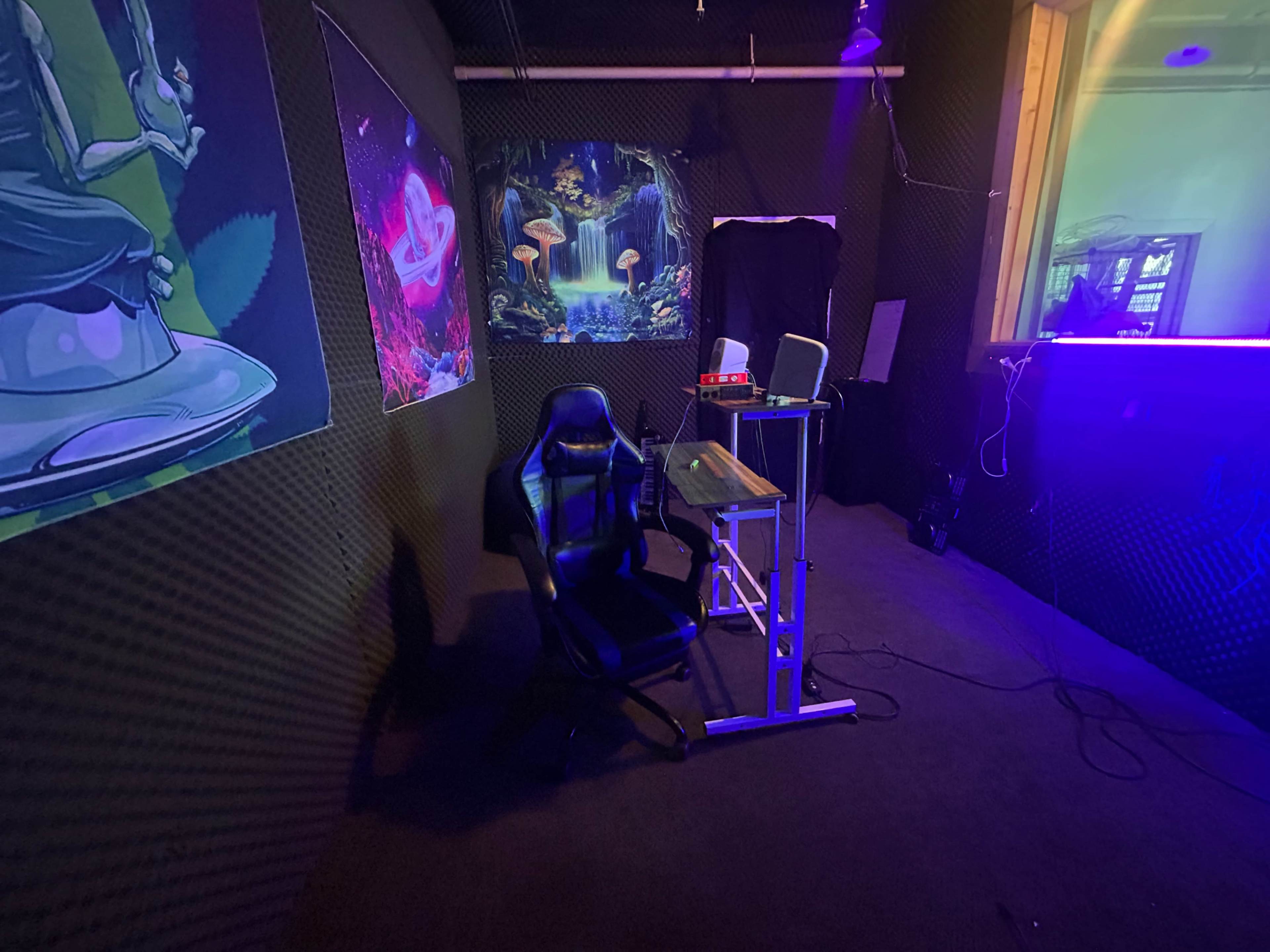 The image shows a dimly lit room with a gaming chair and a desk featuring a projector, decorated with vibrant wall art and neon lighting.