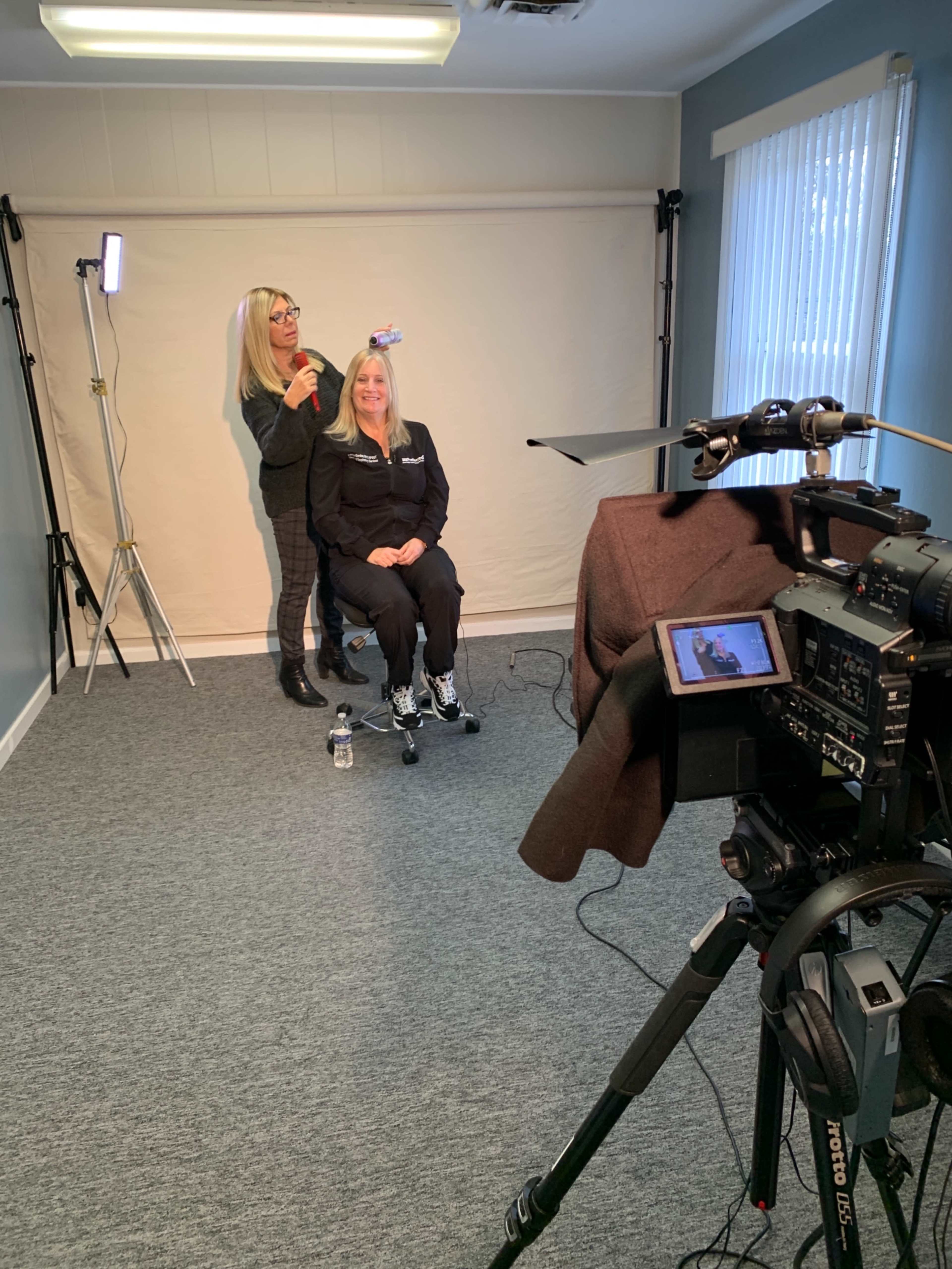 A woman is applying makeup to another woman in a studio setting, with a camera and lighting equipment positioned for a photoshoot.