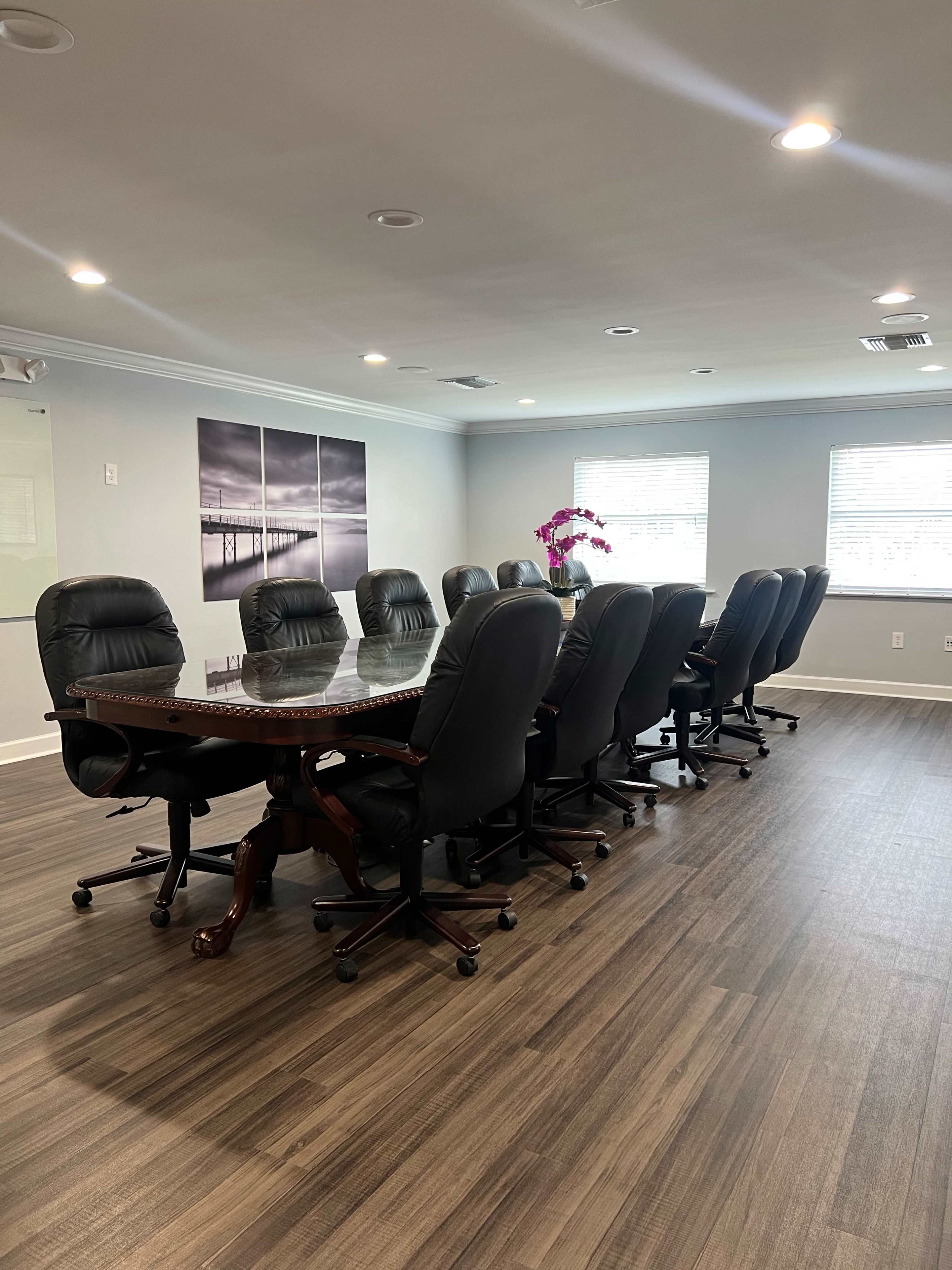 A spacious conference room featuring a large wooden table surrounded by twelve black leather office chairs, with two framed black-and-white photographs on the wall and a potted orchid in the corner.