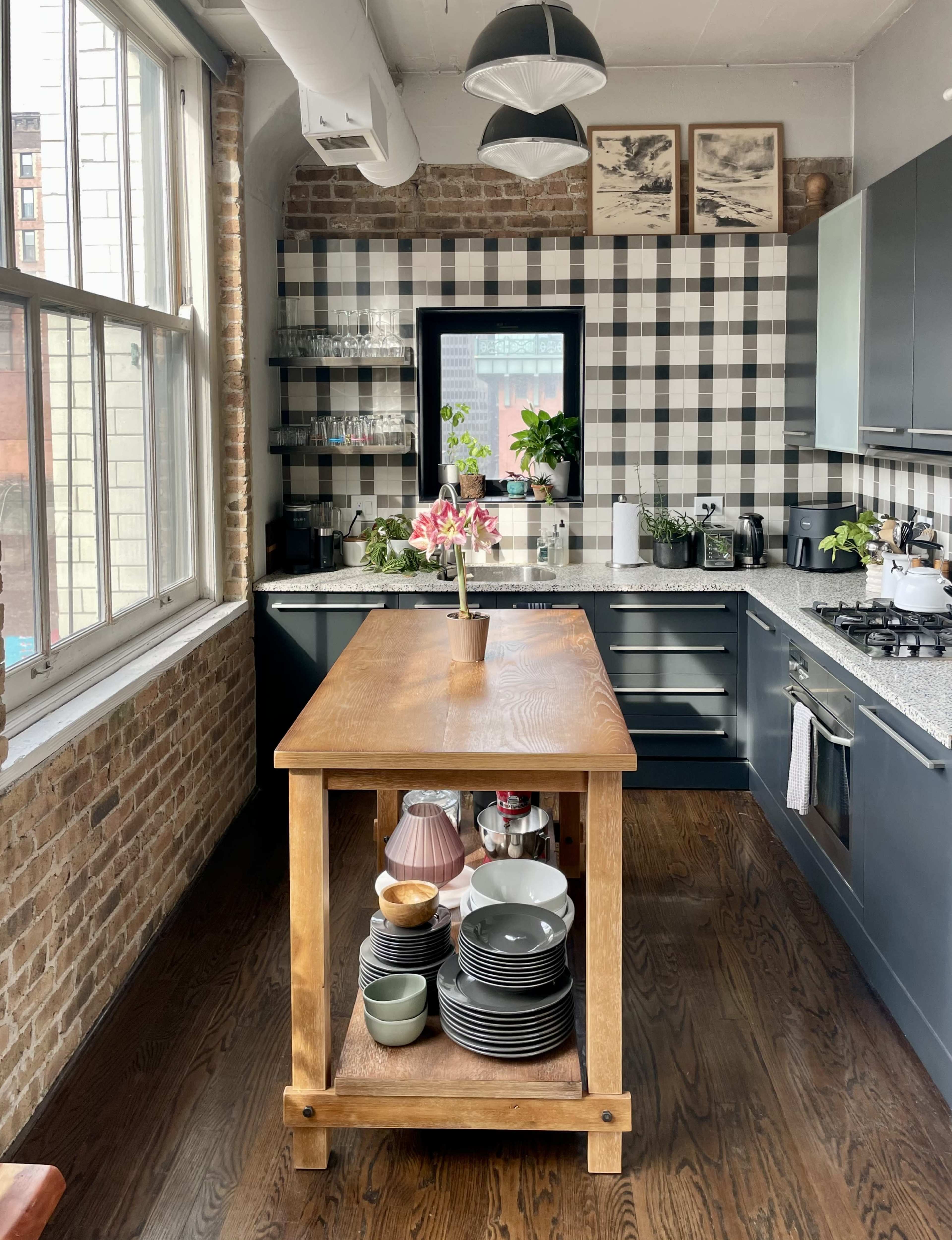 A modern kitchen features a central wooden island, checkered wall design, and various plants alongside dishware on display.