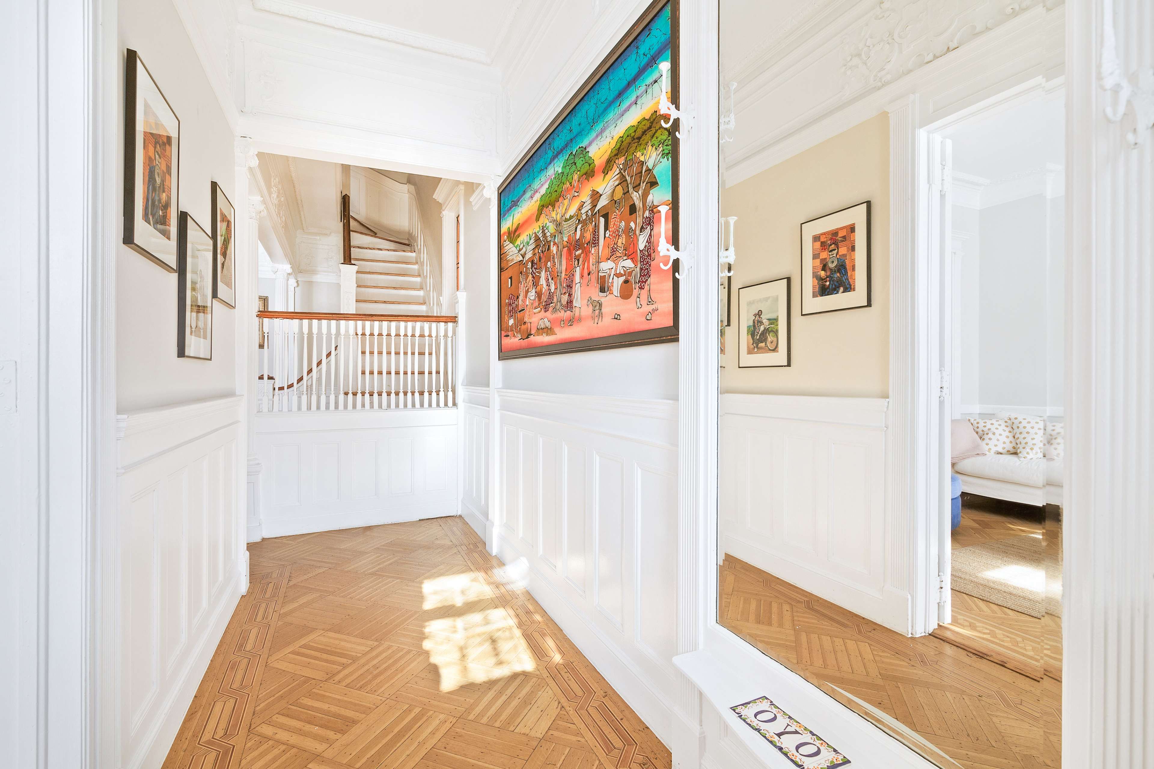 A hallway features a colorful painting on the wall, with stairs visible in the background and light streaming through a window.