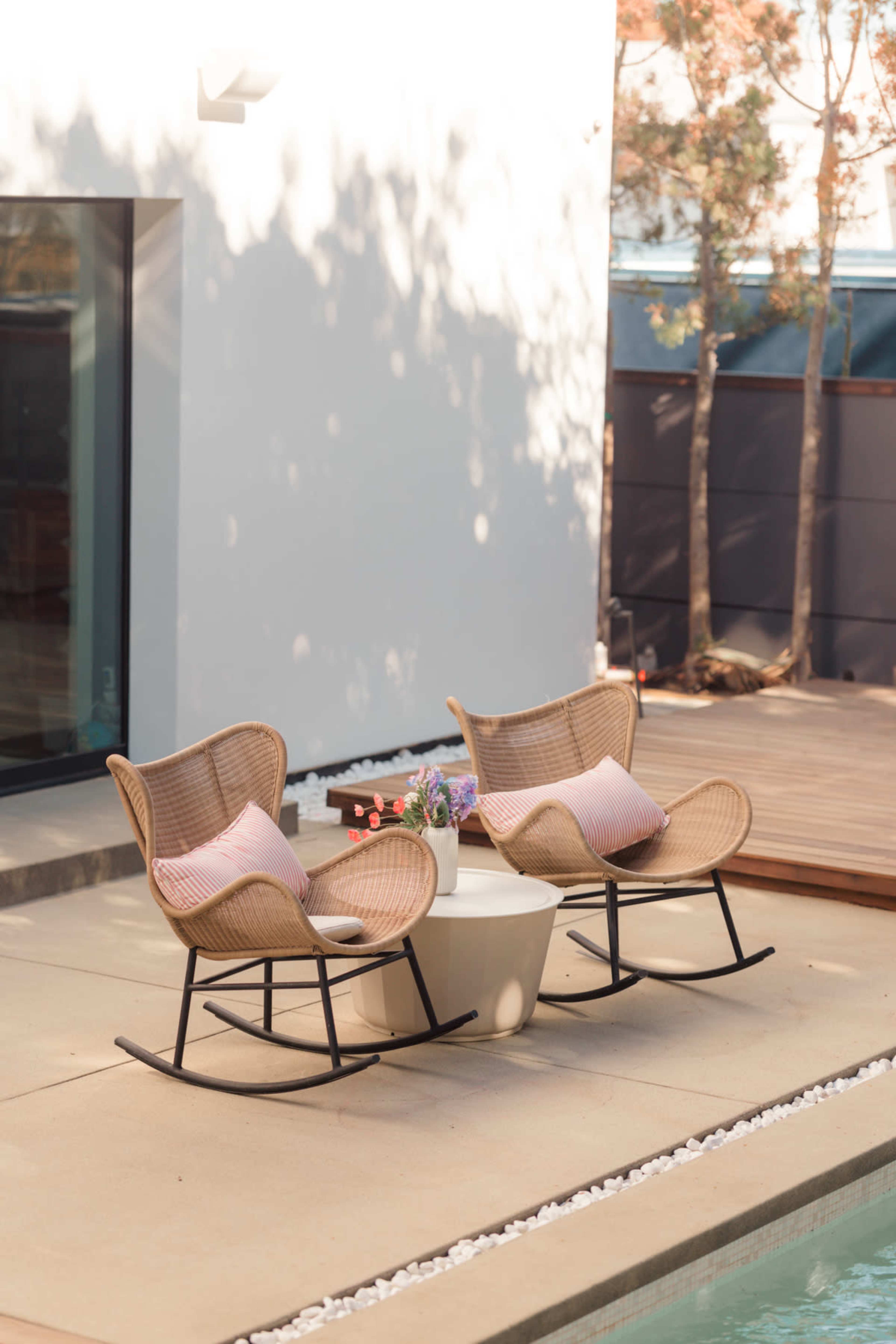 The image shows two wicker rocking chairs with cushions placed beside a swimming pool on a concrete patio.