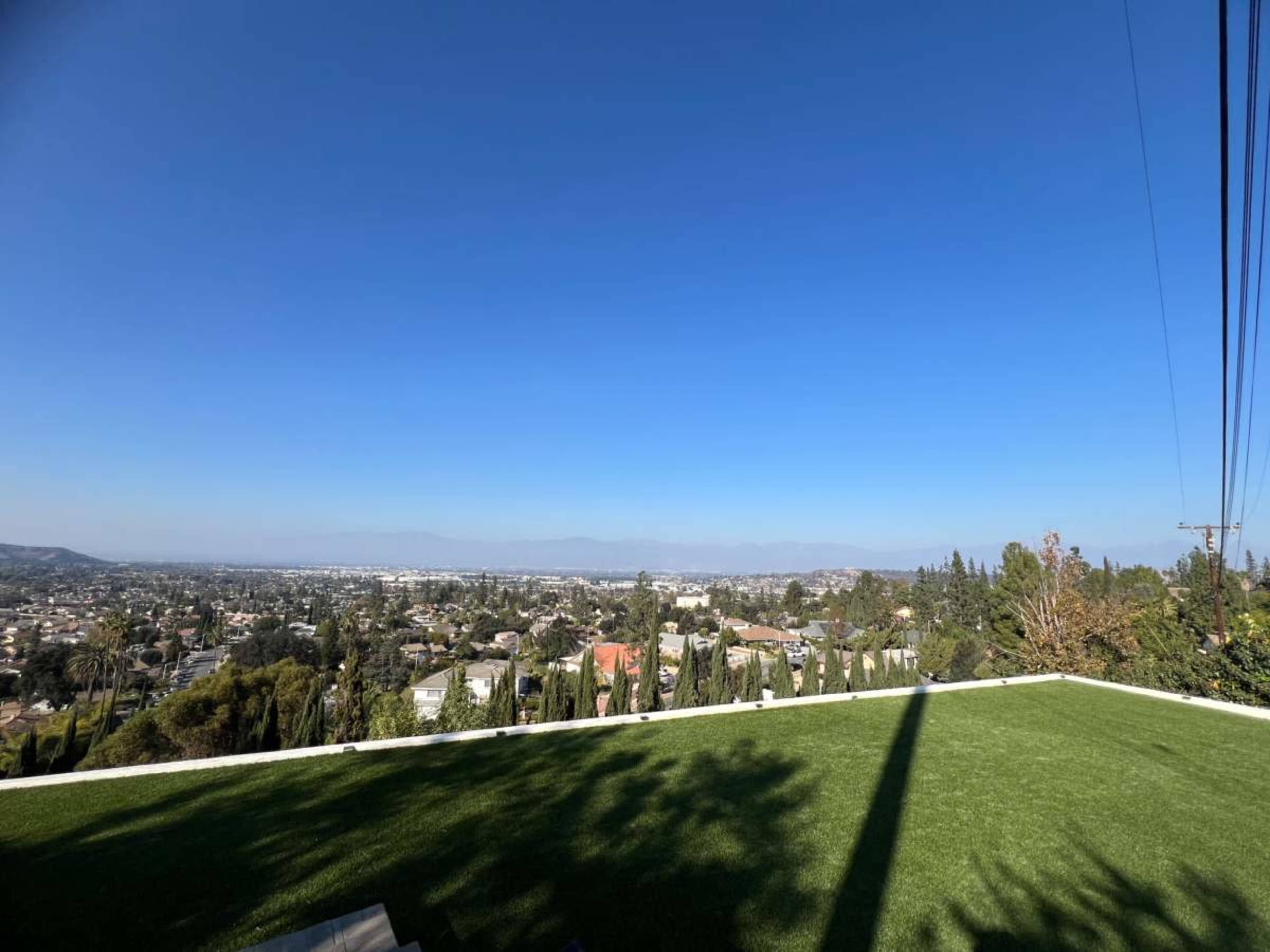 The image shows a scenic view of a city from an elevated grassy area, with clear blue skies and various buildings and trees visible in the distance.