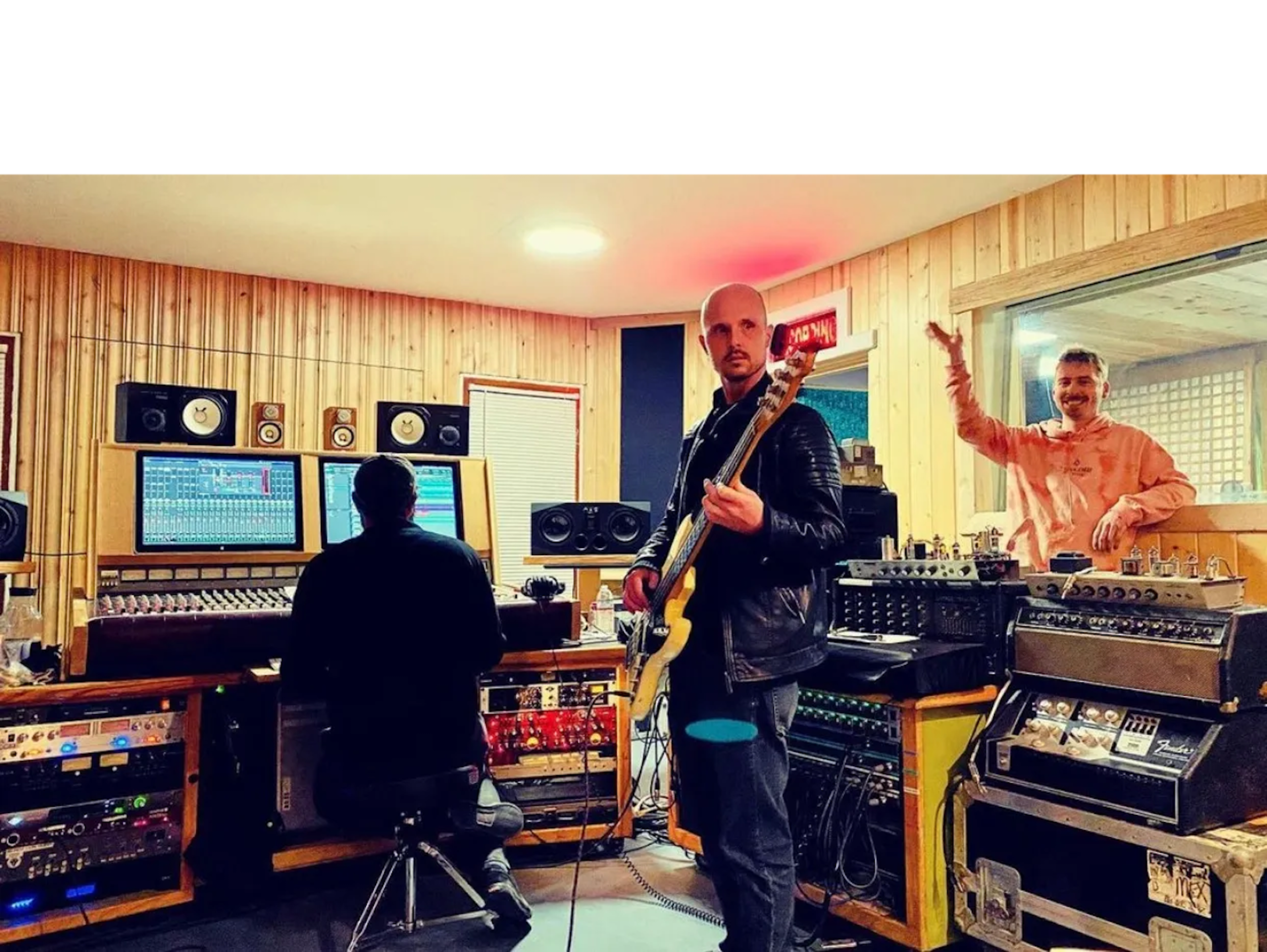 Three musicians are working in a recording studio, with one playing bass and another engineer seated at a mixing console.