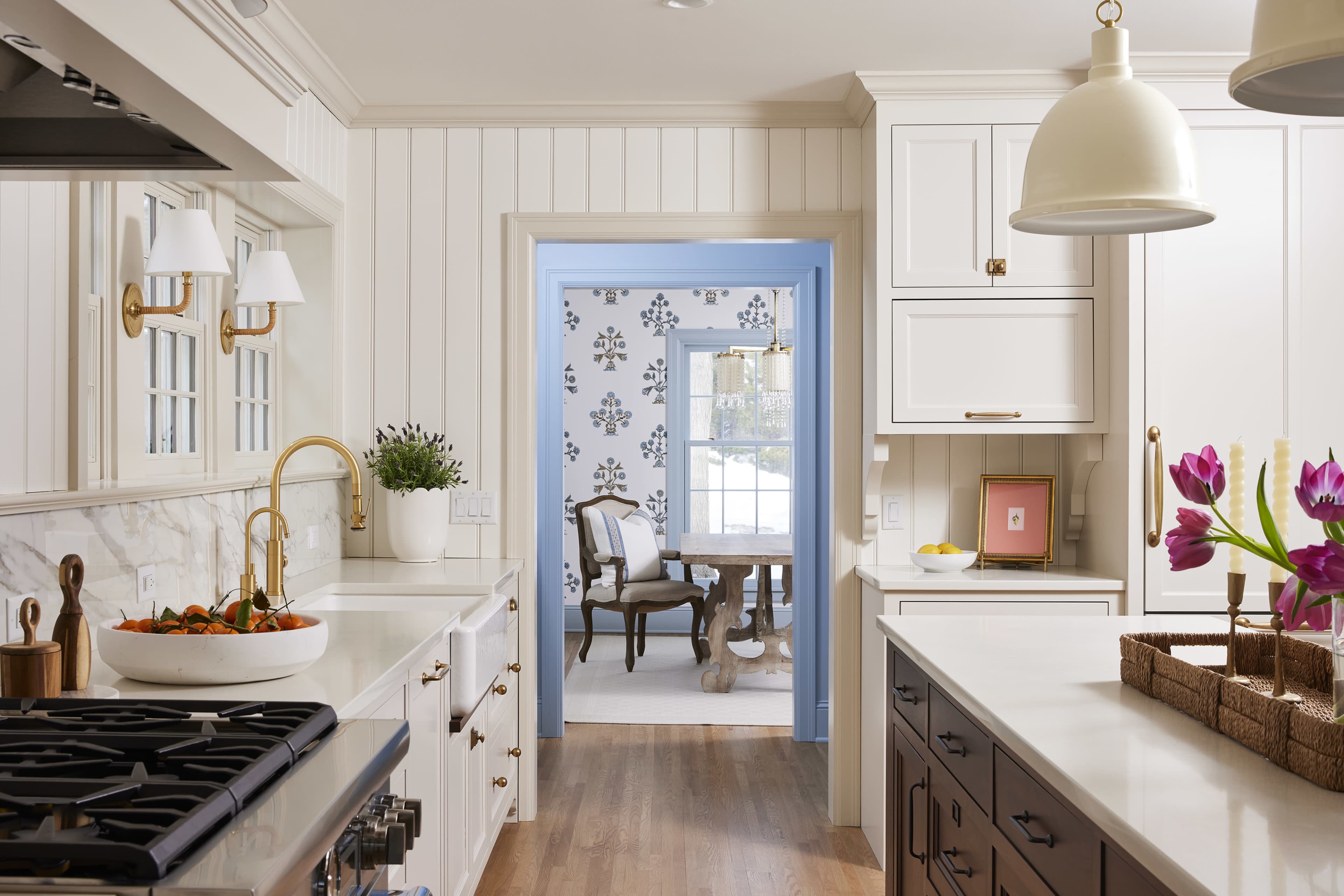 A bright kitchen features white cabinetry, a gas stove, and a view into a dining area with patterned wallpaper and a wooden table.