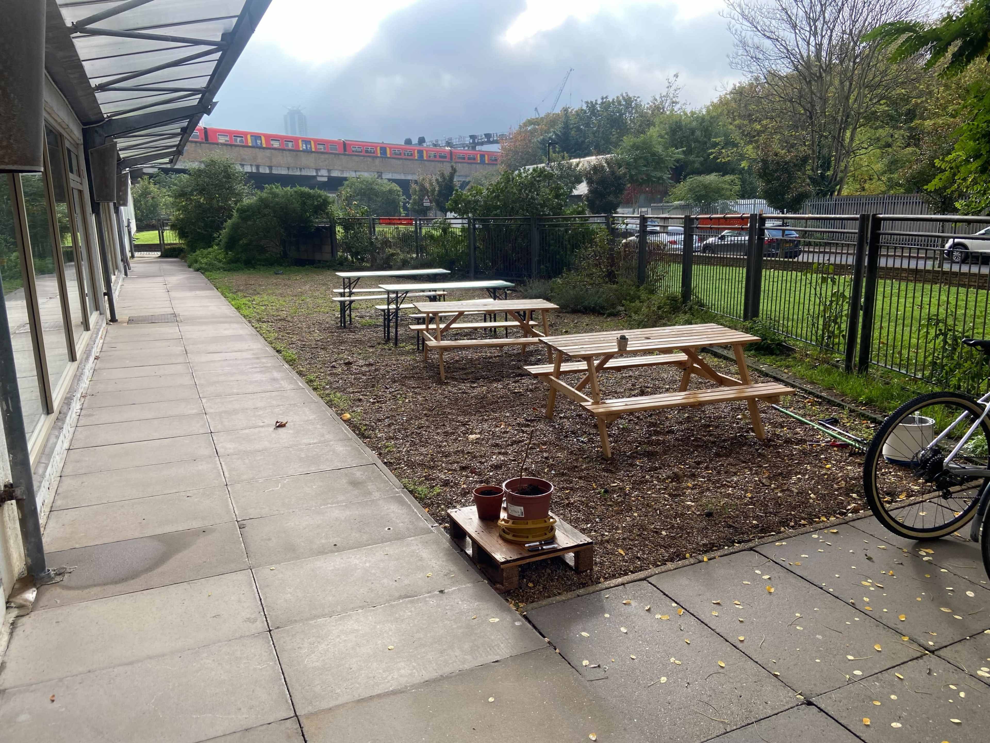 A row of wooden picnic tables is situated on a grassy area next to a paved walkway, with a train visible in the background.