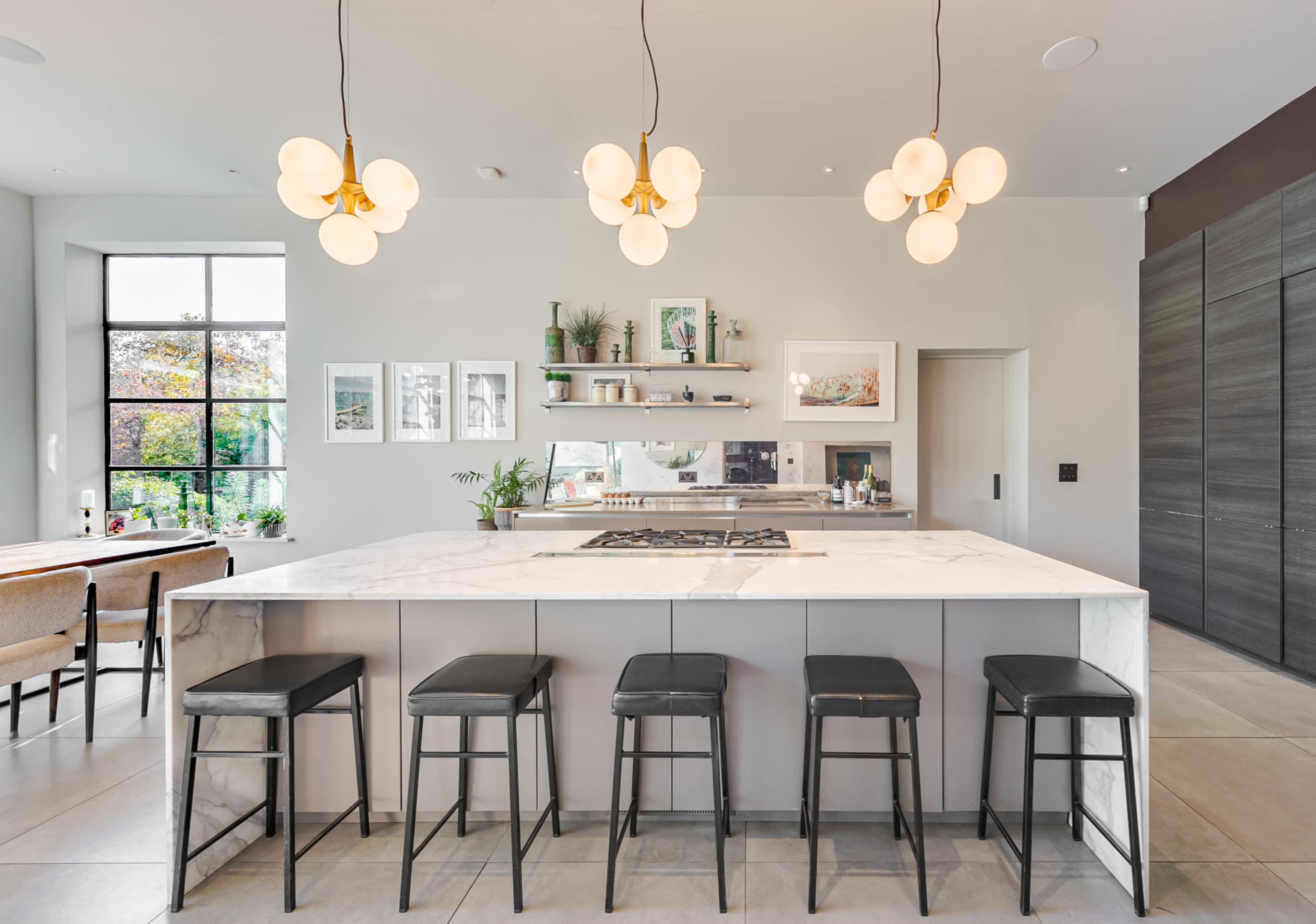 A modern kitchen features a large marble island with black stools, elegant pendant lighting, and shelves displaying decorative items against a light-colored wall.
