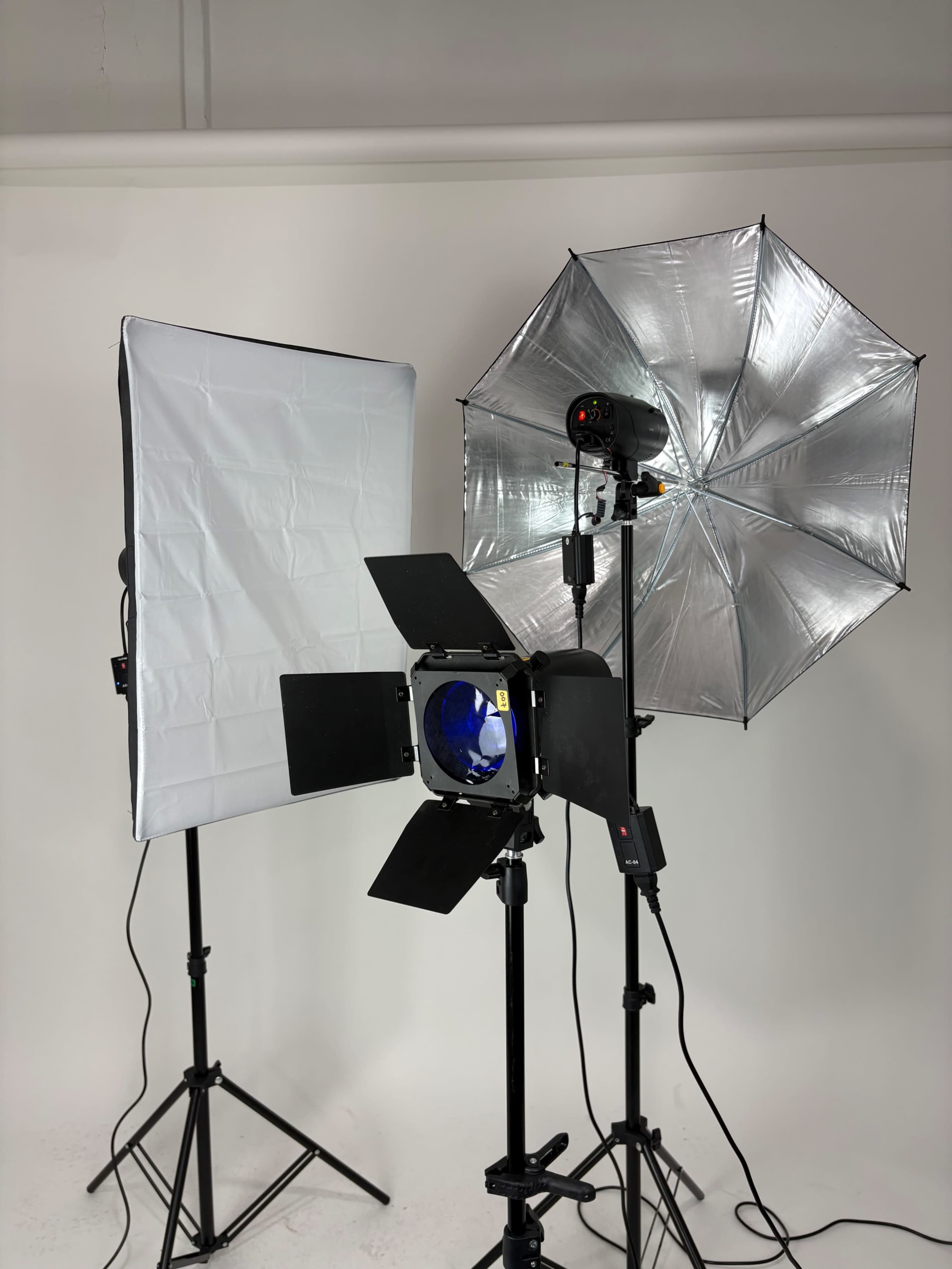 The image shows a photography setup featuring a softbox, an umbrella light, and a spotlight, all on adjustable tripods against a neutral backdrop.