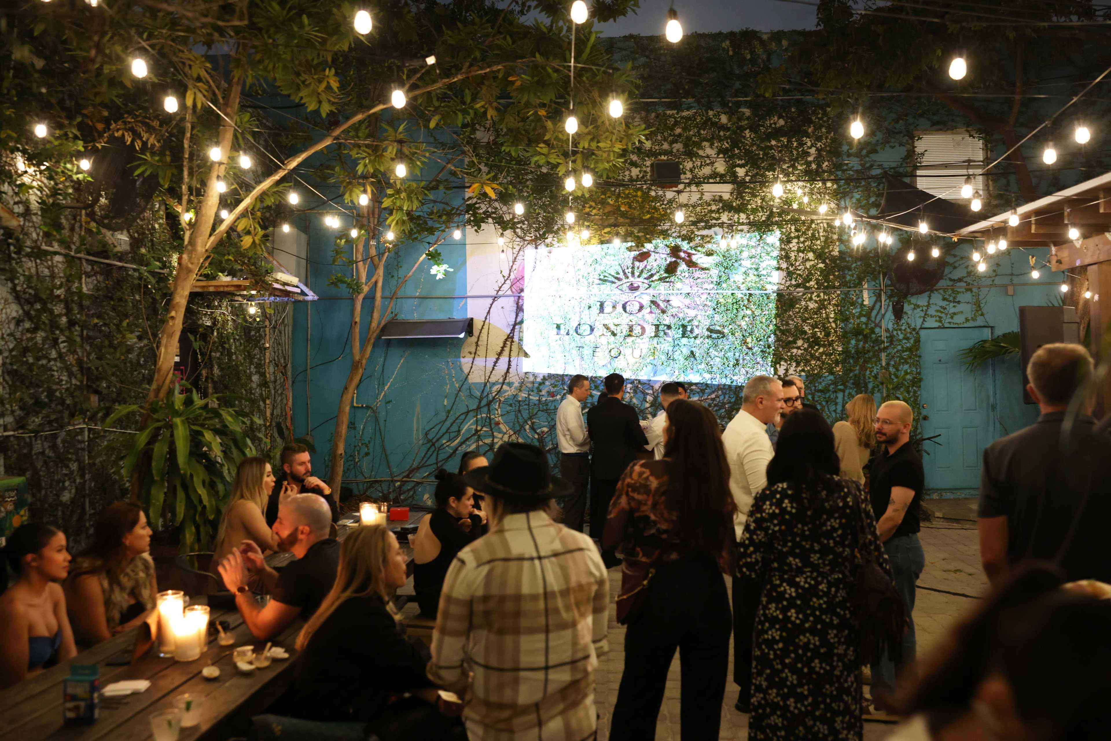A group of people socializes in an outdoor venue decorated with string lights and foliage, featuring a large projected screen displaying a logo.