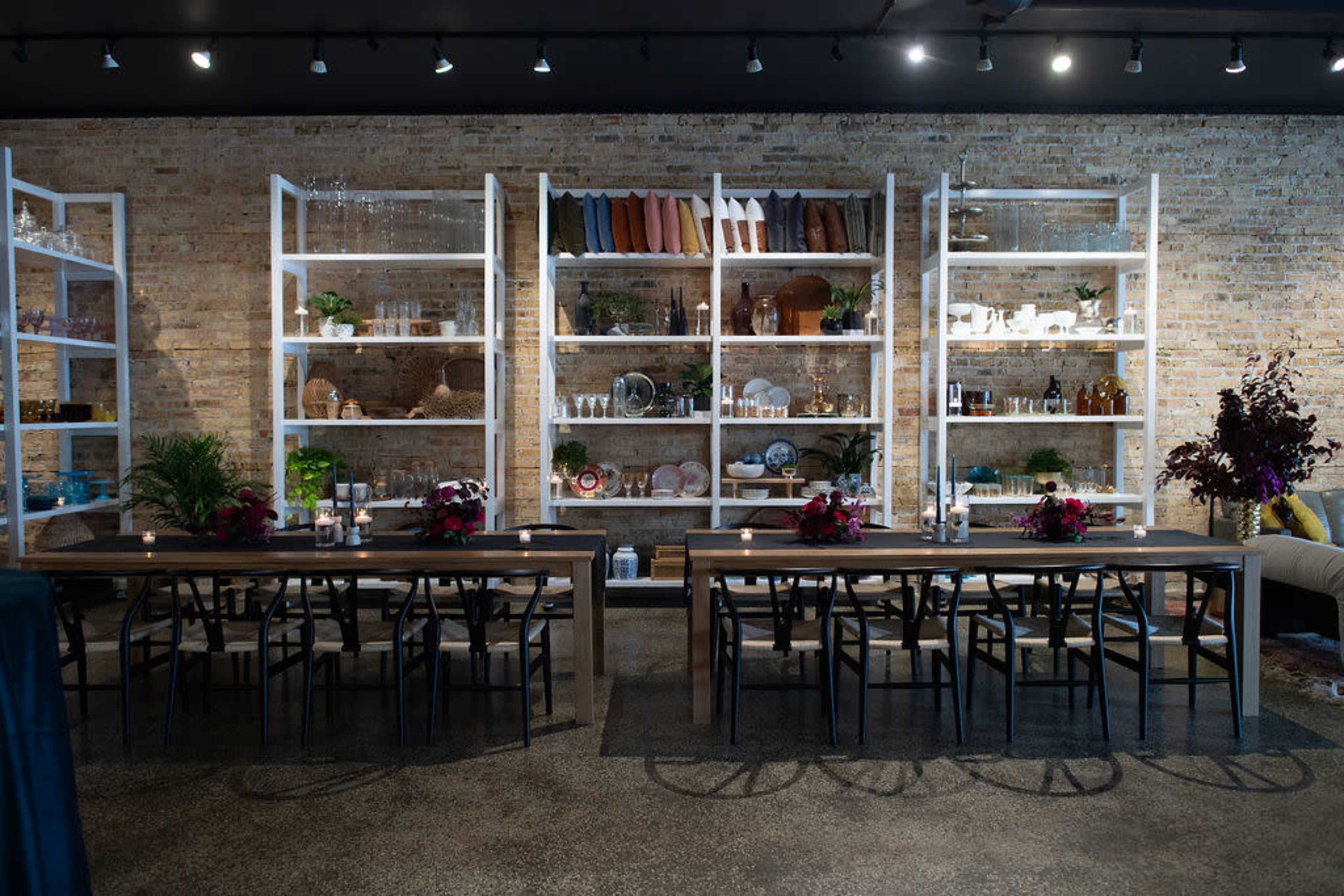 A modern dining area with two long wooden tables and a wall of shelves displaying glassware and decorative items against a brick backdrop.